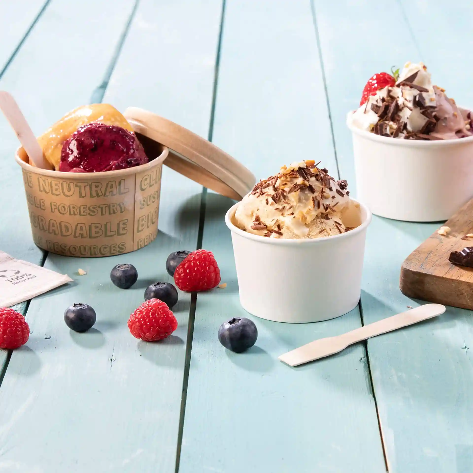 Image de trois coupes de glace avec différentes saveurs, des framboises fraîches, des myrtilles et une cuillère en bois sur une surface en bois bleu.