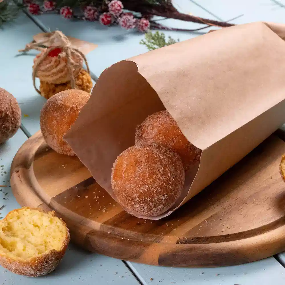 Un sac en papier brun contient plusieurs beignets ronds saupoudrés de sucre glace sur une planche à découper en bois, avec un beignet partiellement coupé.