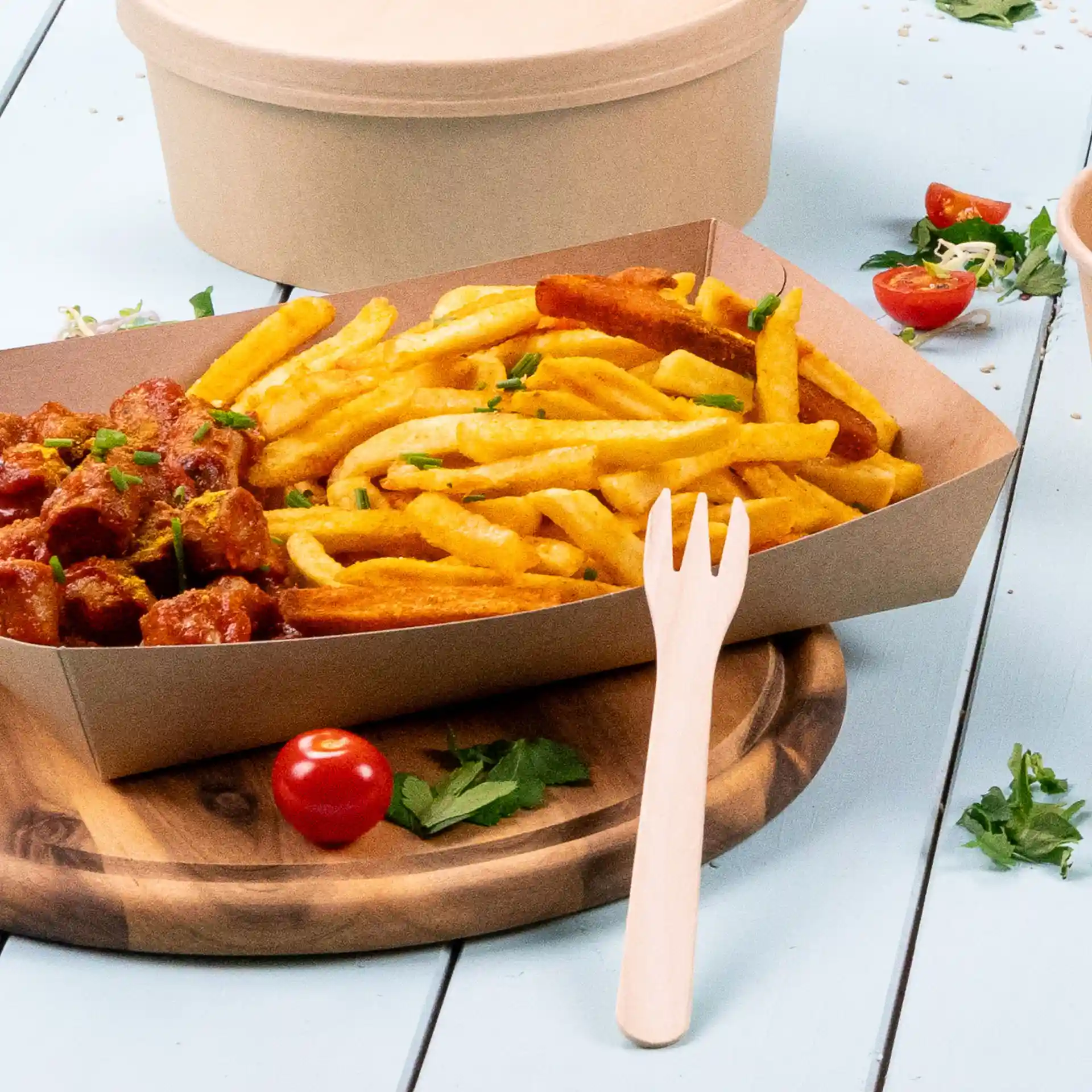 Image d'un plateau en carton rempli de frites et d'un plat de viande, accompagné d'une fourchette en bois et d'une tomate cerise sur une planche en bois.