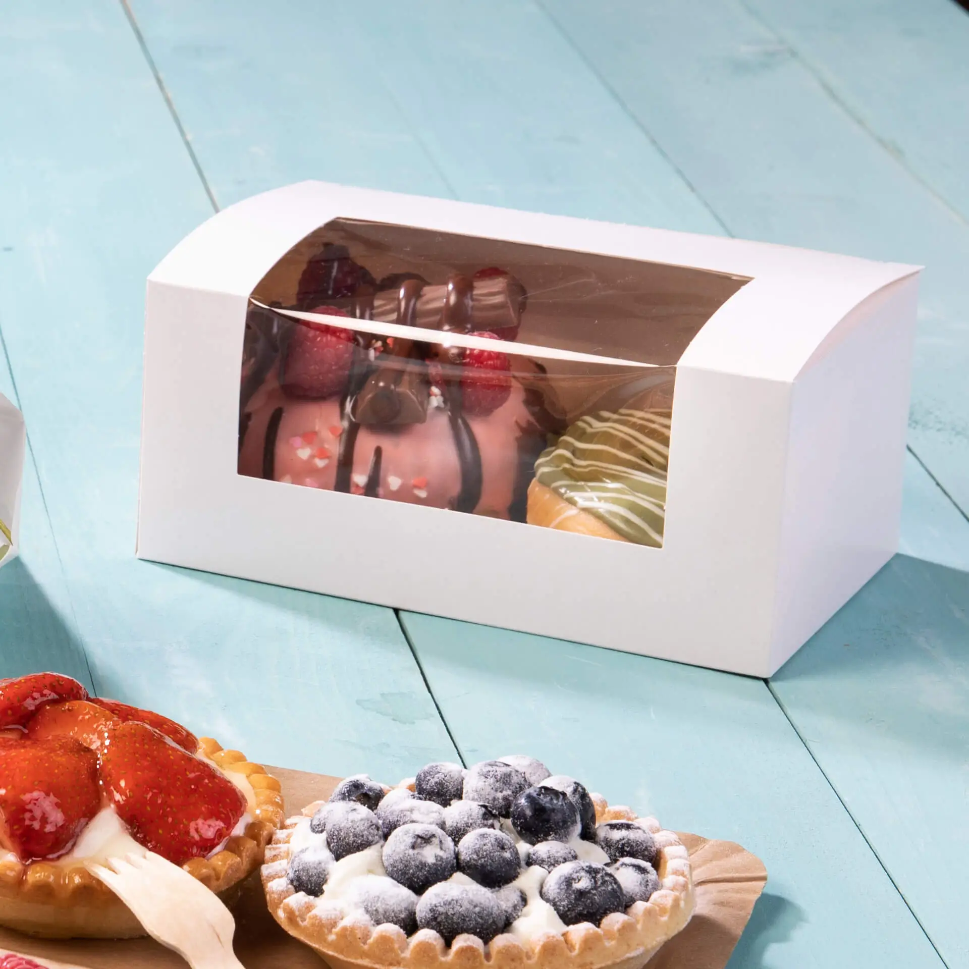 Boîte à cupcakes blanche avec une fenêtre en cellulose affichant des pâtisseries variées, y compris gâteaux et tartes, sur une surface turquoise.