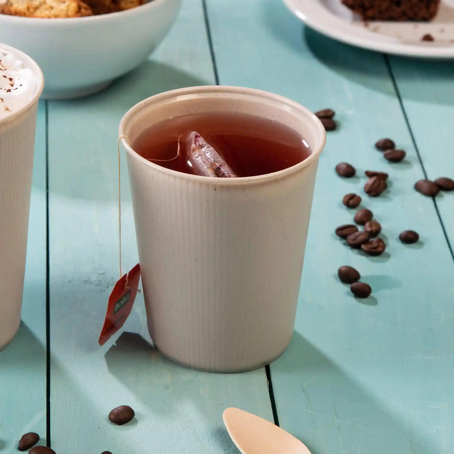 Gobelet réutilisable crème blanc avec sachet de thé à l'intérieur, posé sur une surface en bois bleu avec des grains de café à proximité.