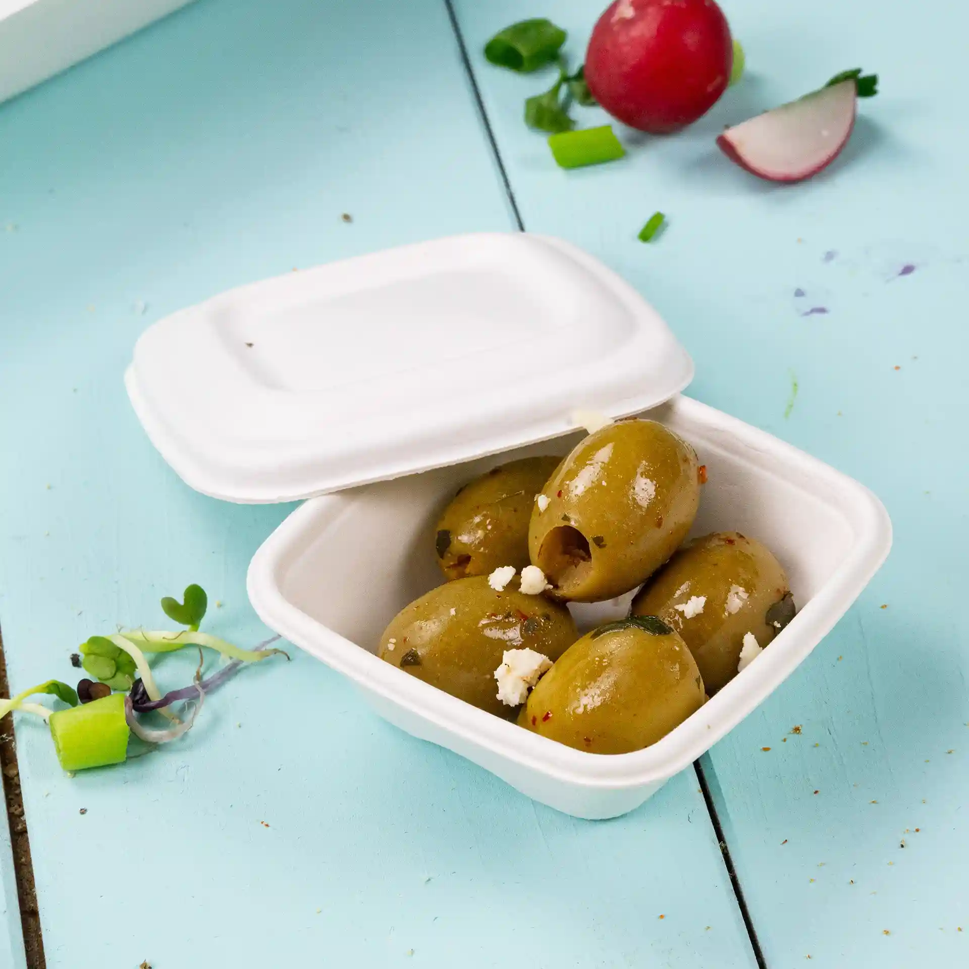 Gobelet carré blanc contenant plusieurs olives vertes, placé sur une surface bleu clair avec des légumes éparpillés.