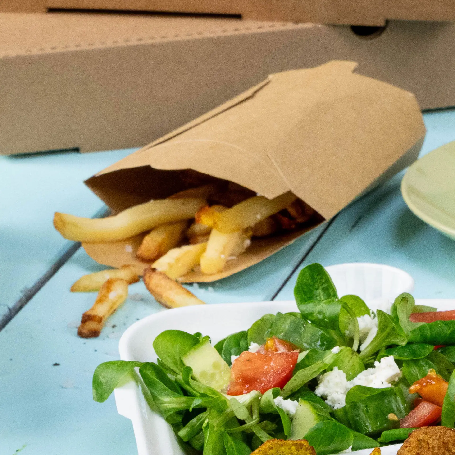 Une pelle en kraft avec des frites débordantes ; une salade fraîche dans un contenant blanc est à proximité sur une surface bleue.