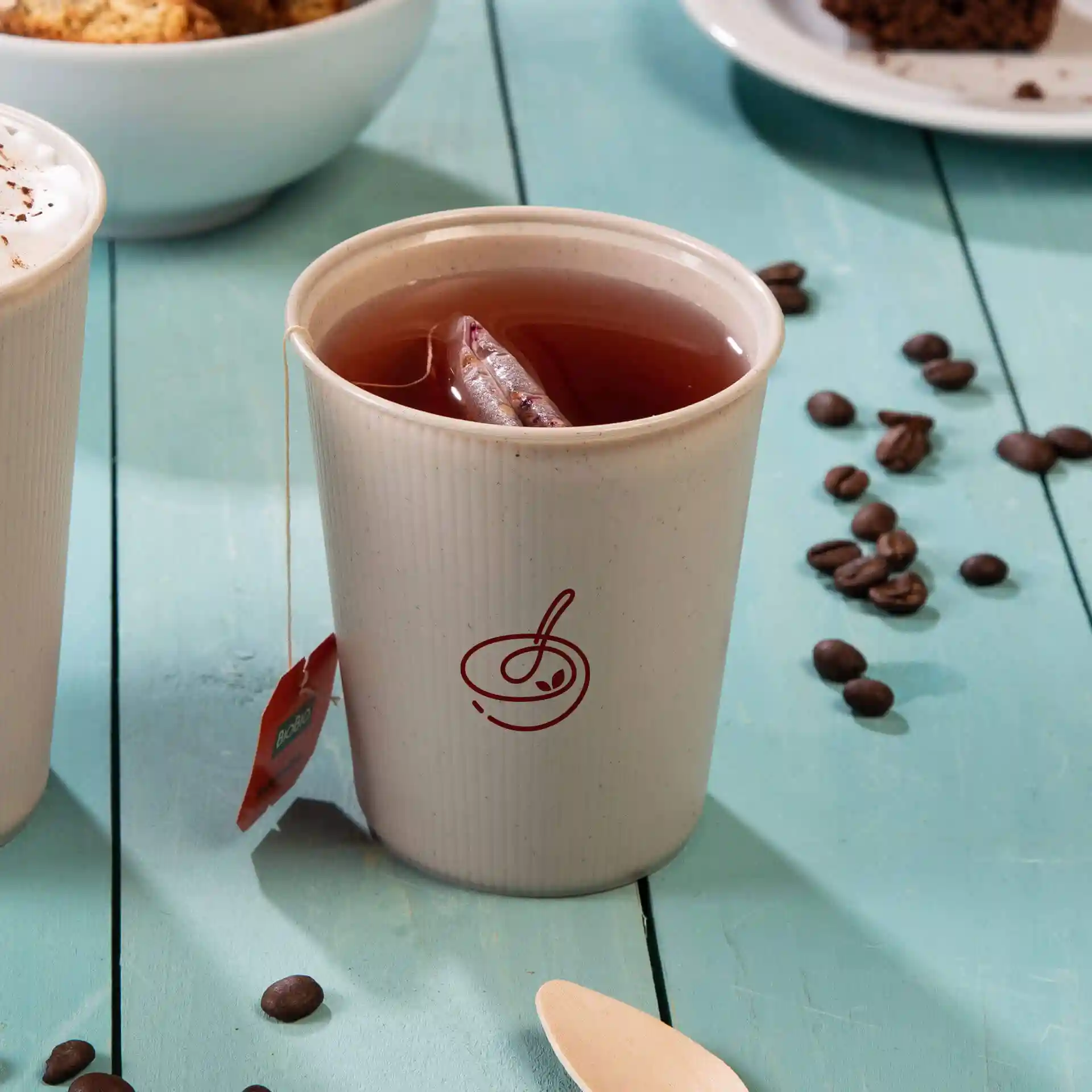Tasse réutilisable blanche avec un design ondulé, remplie de thé et d'un sachet de thé, sur une surface en bois bleu avec des grains de café.