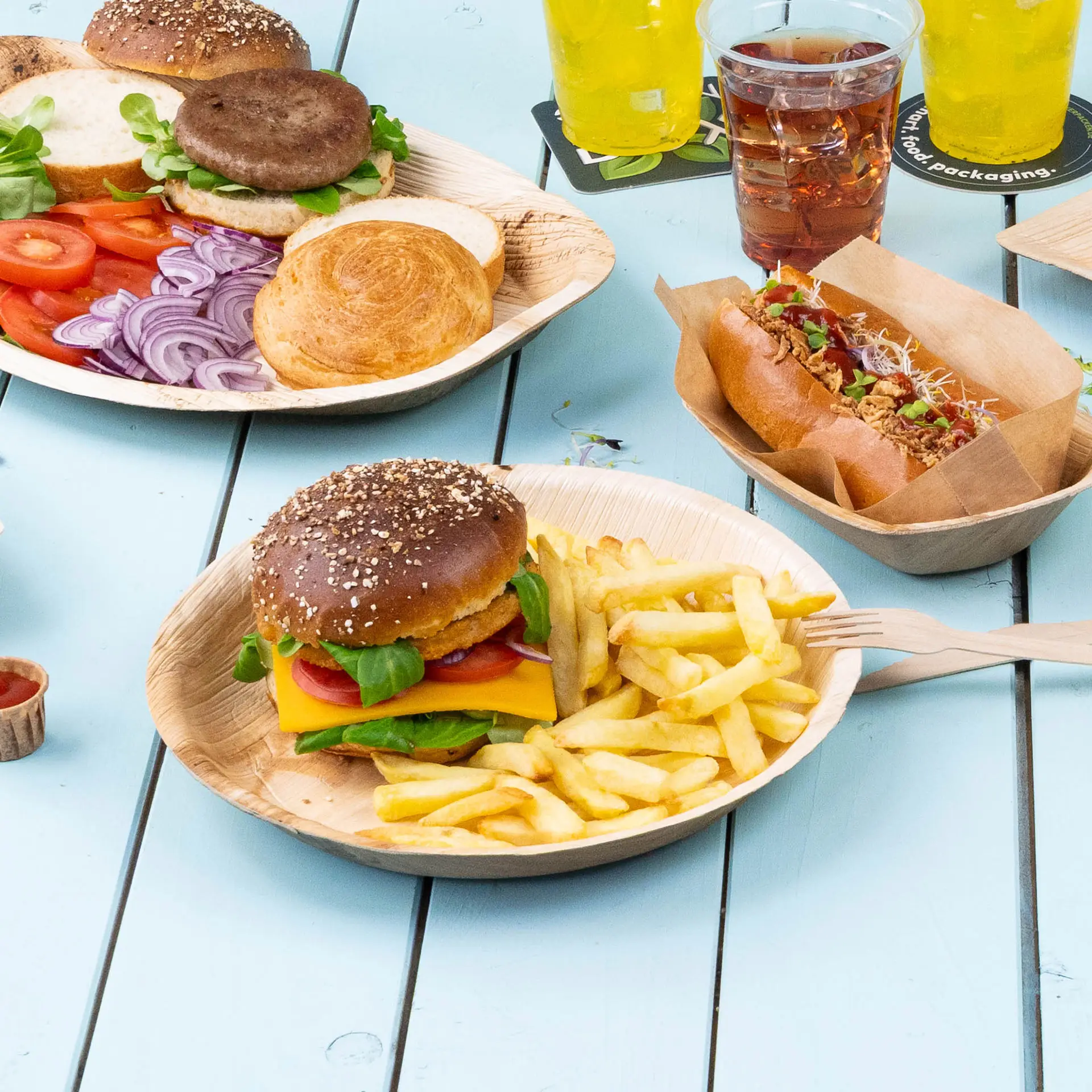 Une assiette en feuille de palmier contient un cheeseburger avec de la laitue, des tomates et des frites, accompagné de tranches de légumes et d'un hot-dog.