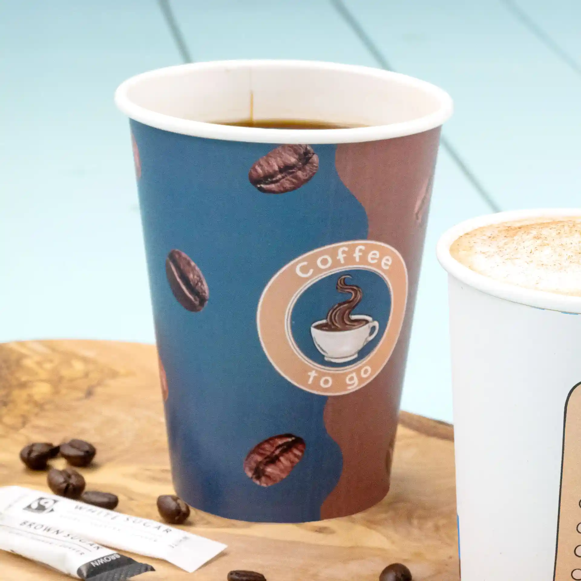 Gobelet en papier avec design imprimé, rempli de café, posé sur une surface en bois avec des sachets de sucre et des grains de café.