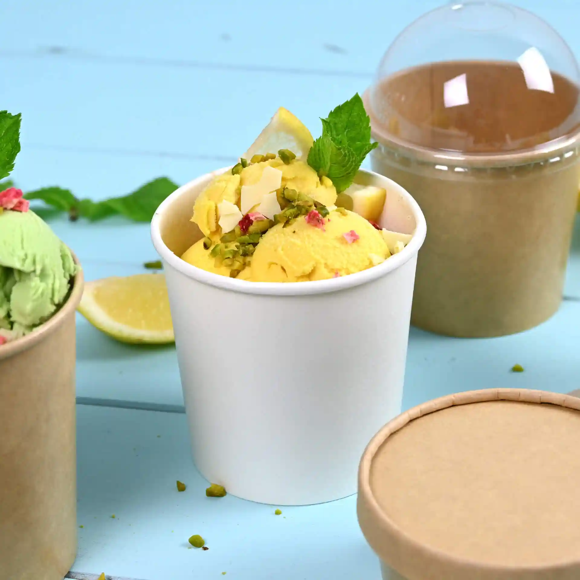Tasse à glace en papier blanc remplie de glace jaune, garnie de feuilles de menthe et de tranches de citron, avec un récipient marron derrière.