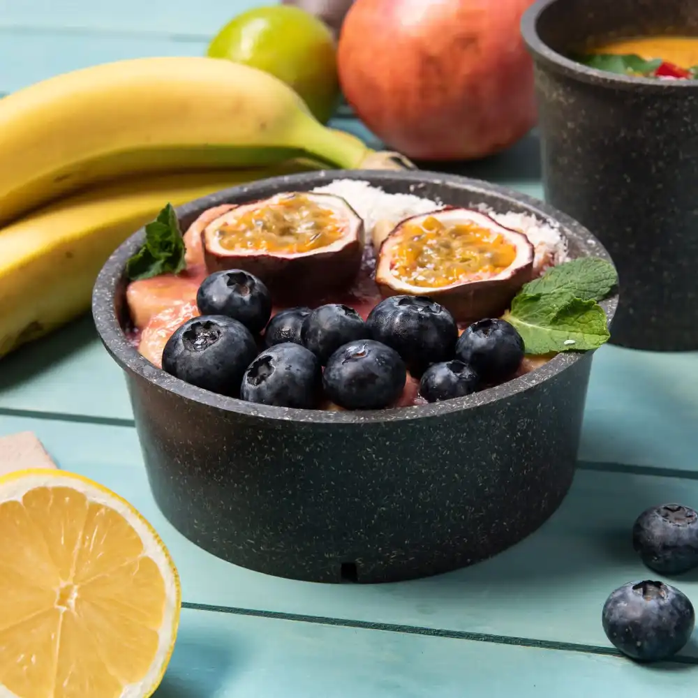 Un bol noir rempli de fruits, y compris des myrtilles et un fruit de la passion, entouré de bananes et d'une tranche de citron.