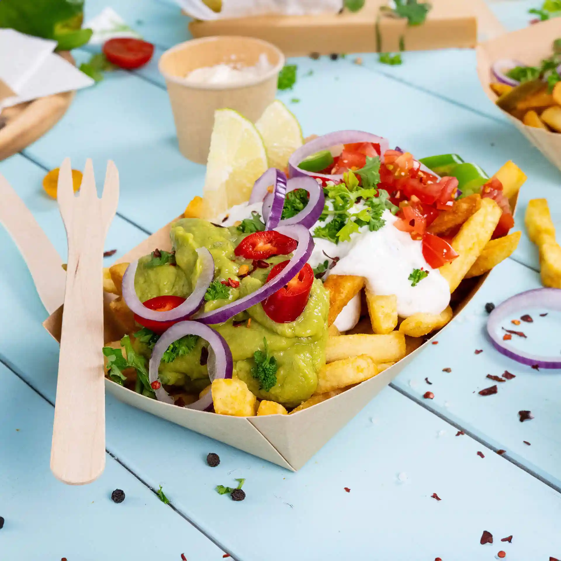 Bol en carton profond rempli de frites, guacamole, crème aigre, tomates en dés, garni d'oignons et de citron vert.