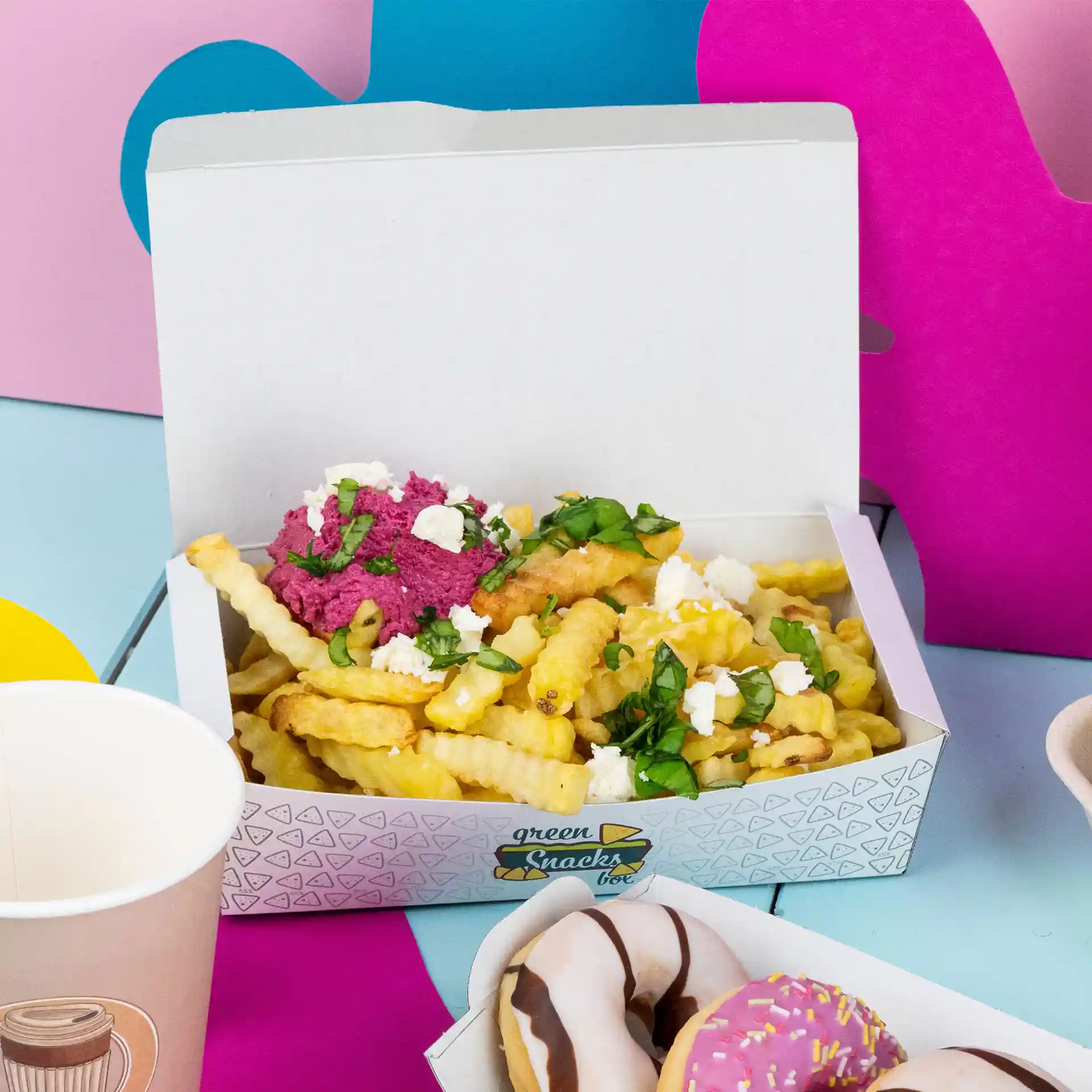 Boîte à snacks blanche contenant des frites ondulées garnies de dip rose, d'herbes et de fromage, avec des donuts et un gobelet à proximité.