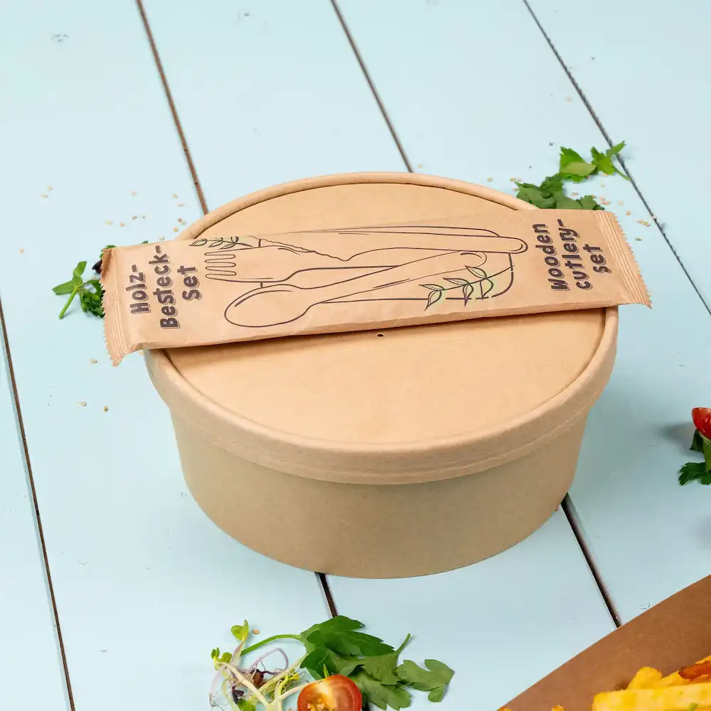 Conteneur brun avec un couvercle et un ensemble de couverts en bois emballé dessus, avec des herbes vertes et des éléments de salade à proximité.