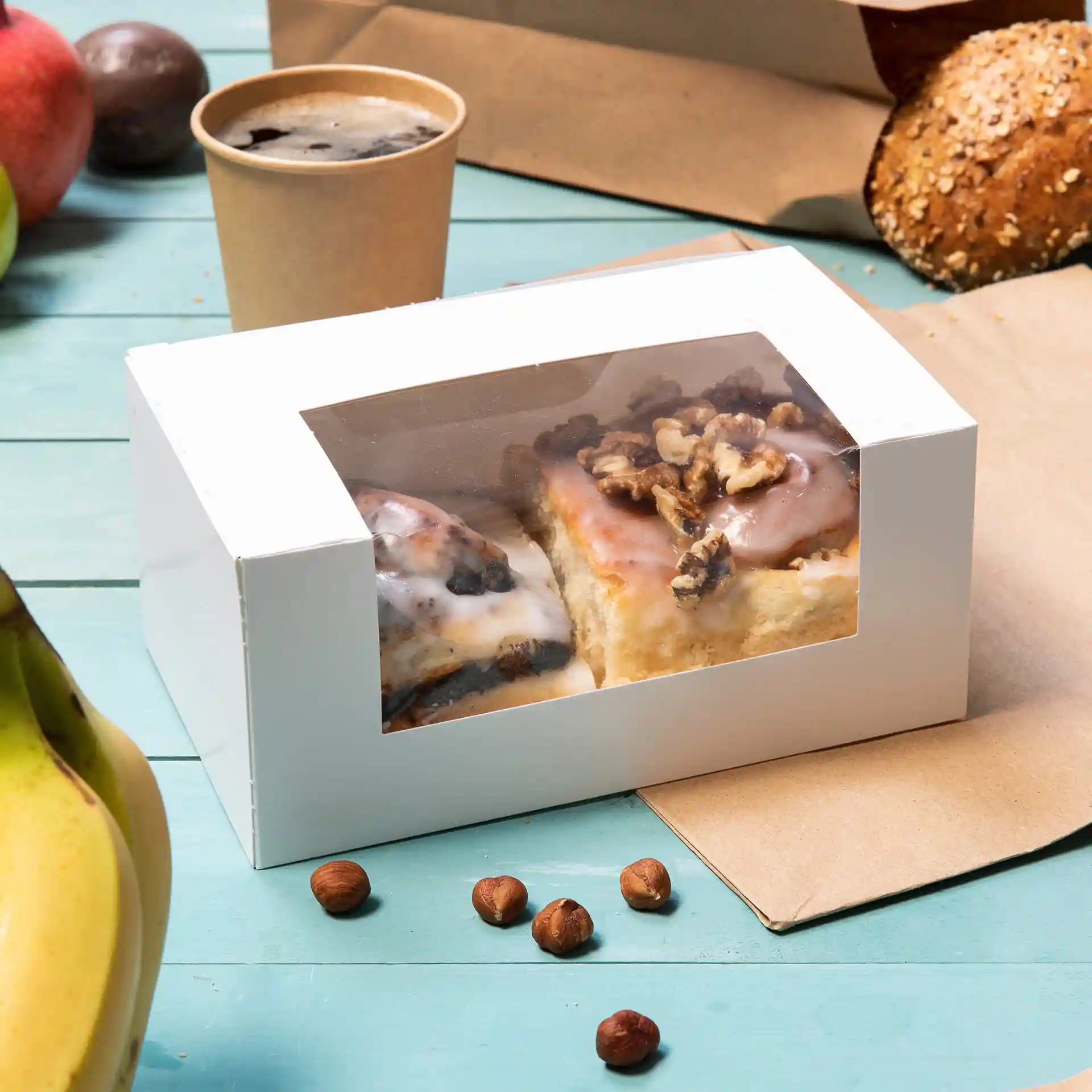 Boîte à pâtisserie blanche avec une fenêtre en cellulose contenant deux pâtisseries, entourée d'une banane, de noisettes et d'une tasse.