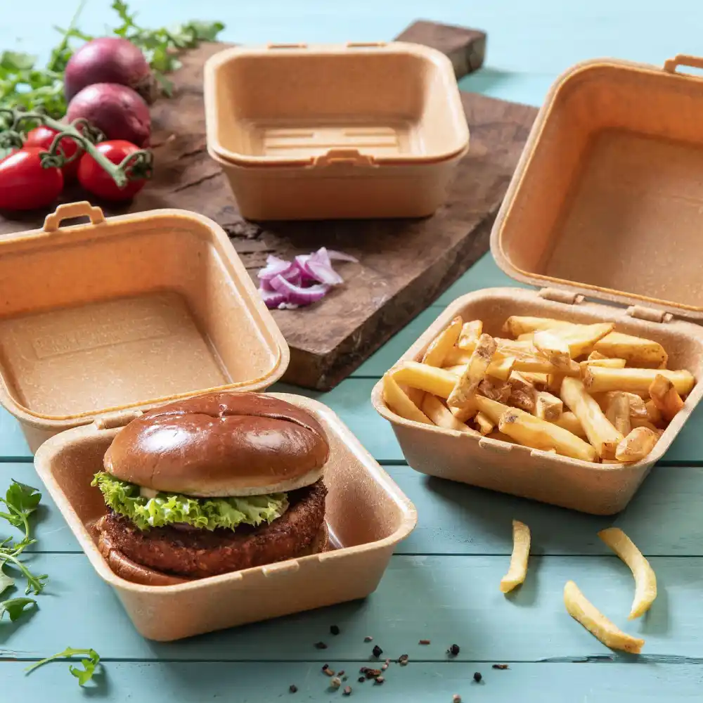 Conteneur avec un burger garni de laitue, à côté d'un conteneur avec des frites, sur une surface en bois avec des légumes à proximité.