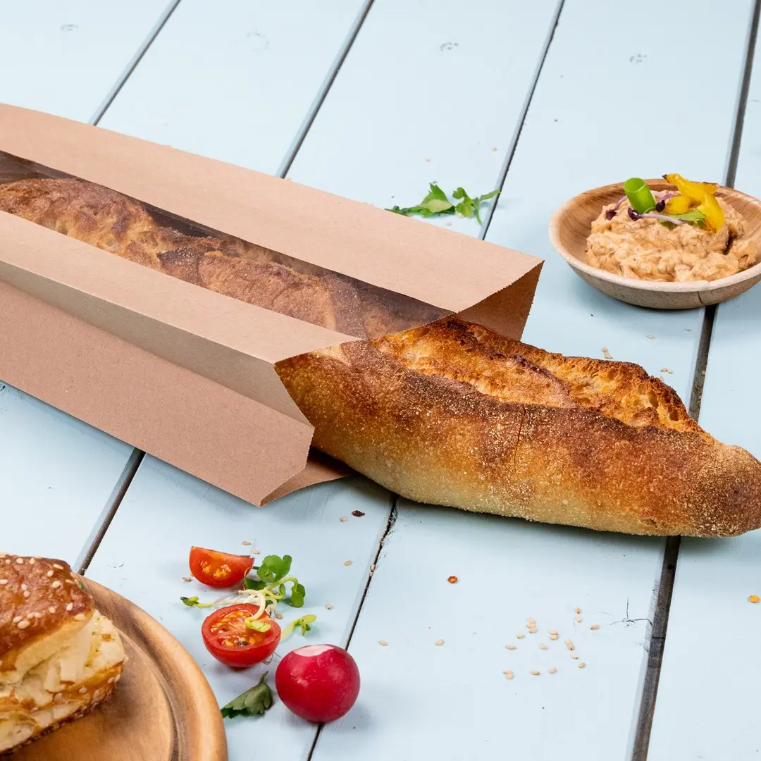 Sac en papier brun partiellement ouvert avec une baguette, des tomates cerises, des radis et un bol de dip sur une surface bleu clair.