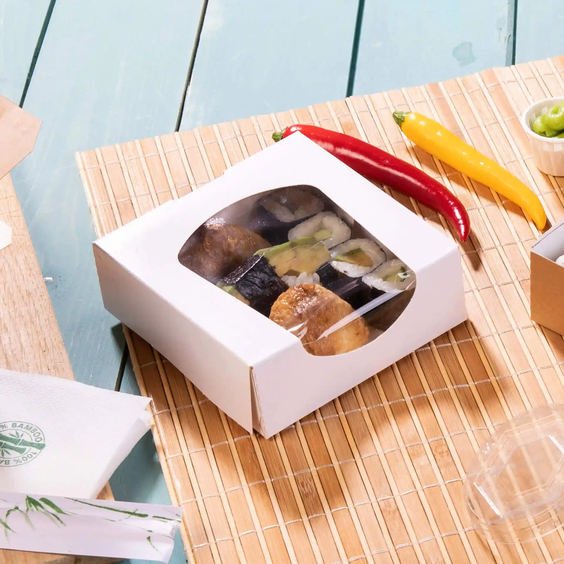 Boîte en carton blanc avec fenêtre contenant des sushis, placée sur un tapis en bambou avec des piments rouges et jaunes à proximité.