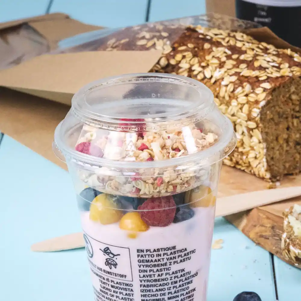 Gobelet avec du yaourt, du granola et des baies, à côté d'un pain à l'avoine sur une surface bleue.