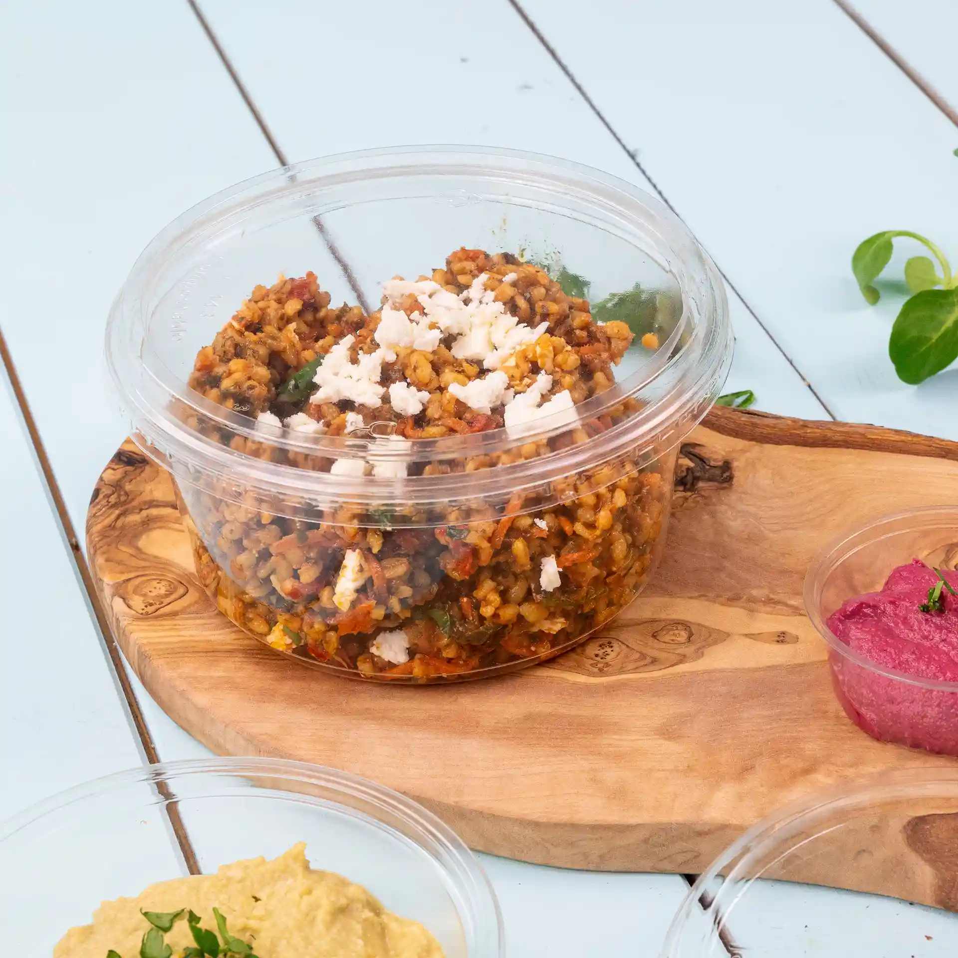 Conteneur rond transparent avec couvercle, rempli d'un plat à base de céréales et garni de fromage émietté, sur une planche en bois.