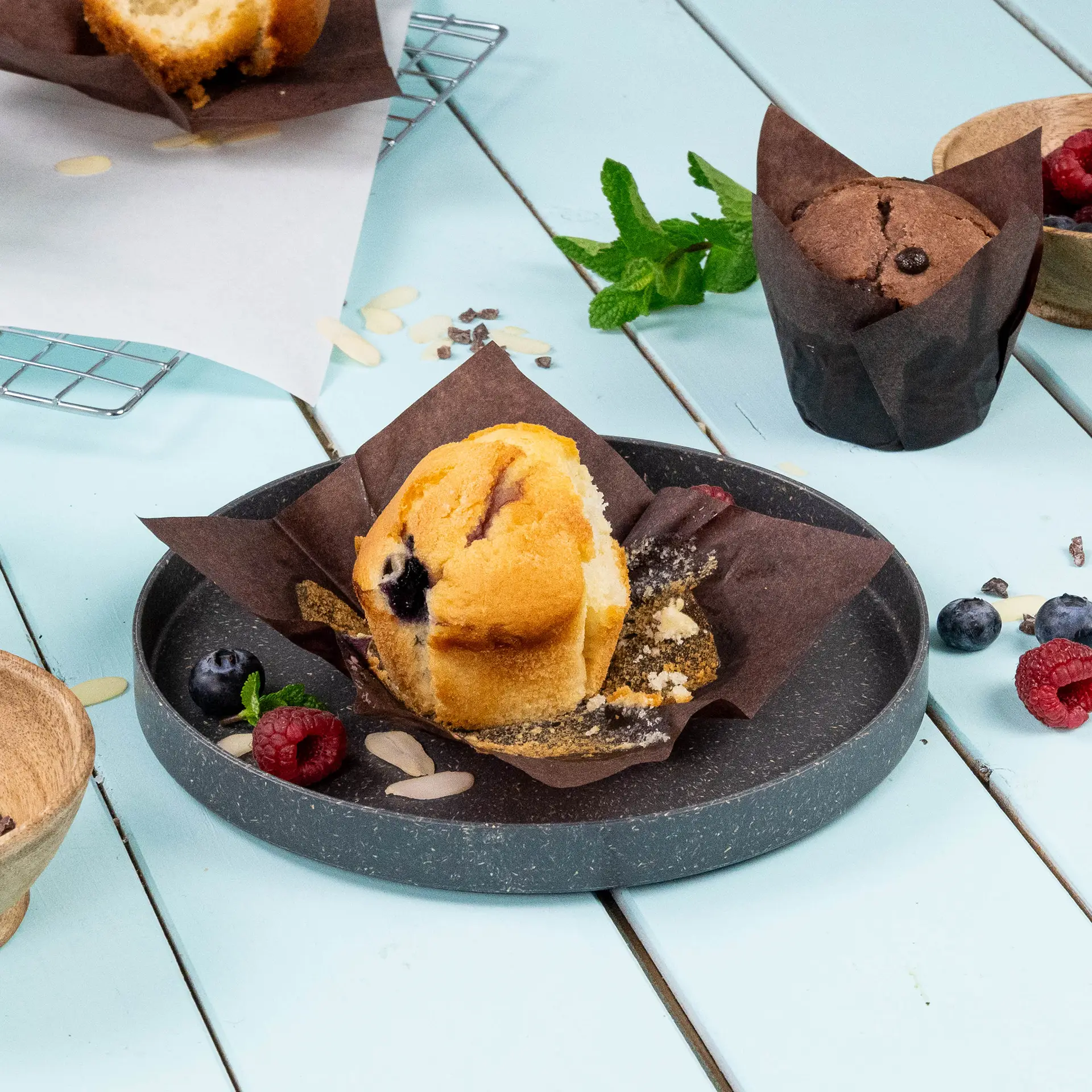 Muffin dans un gobelet en papier brun résistant à la graisse, avec des myrtilles et des framboises sur une assiette grise, sur fond bleu clair.