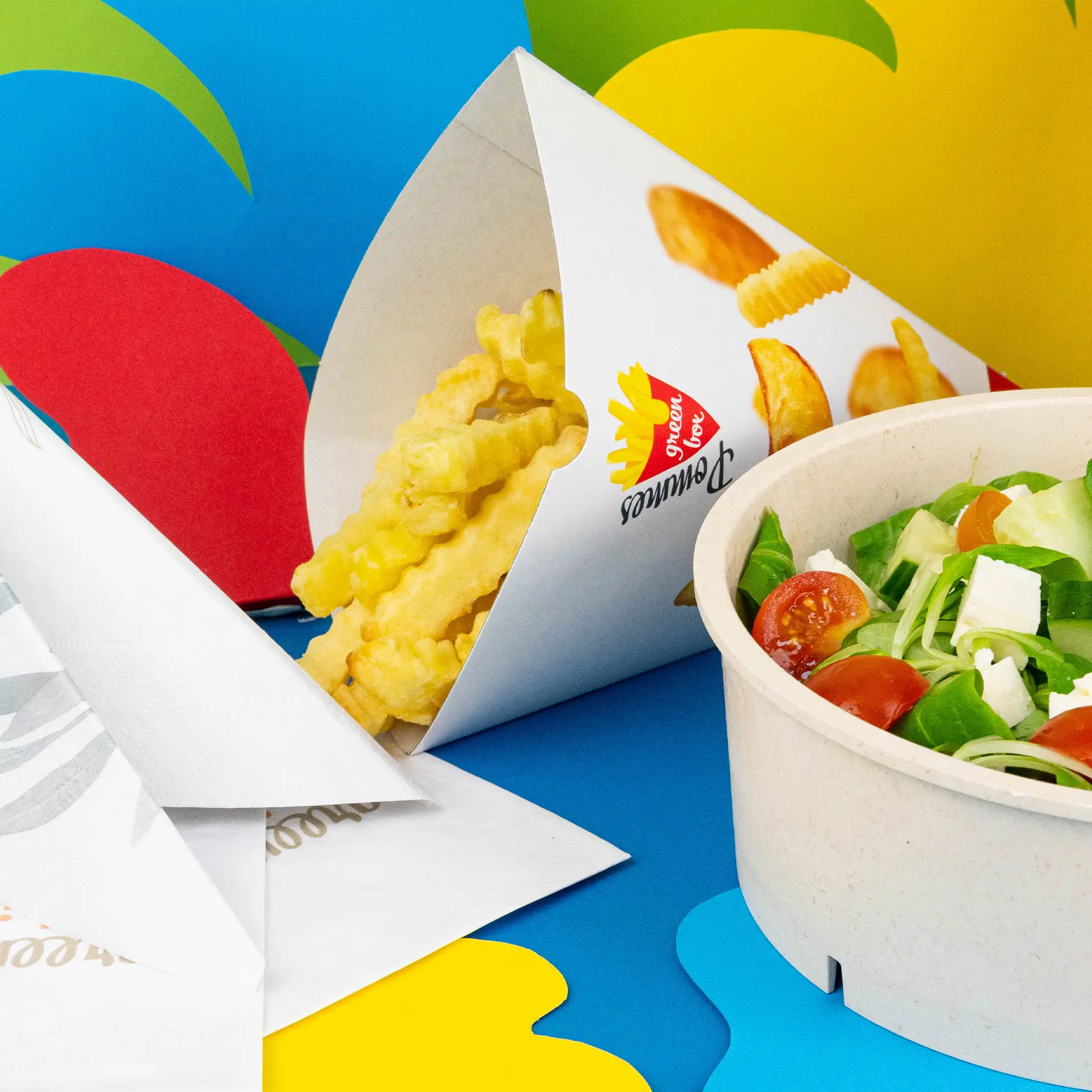 Un cône à snacks imprimé avec des frites ondulées et une salade dans un bol sur un fond coloré.