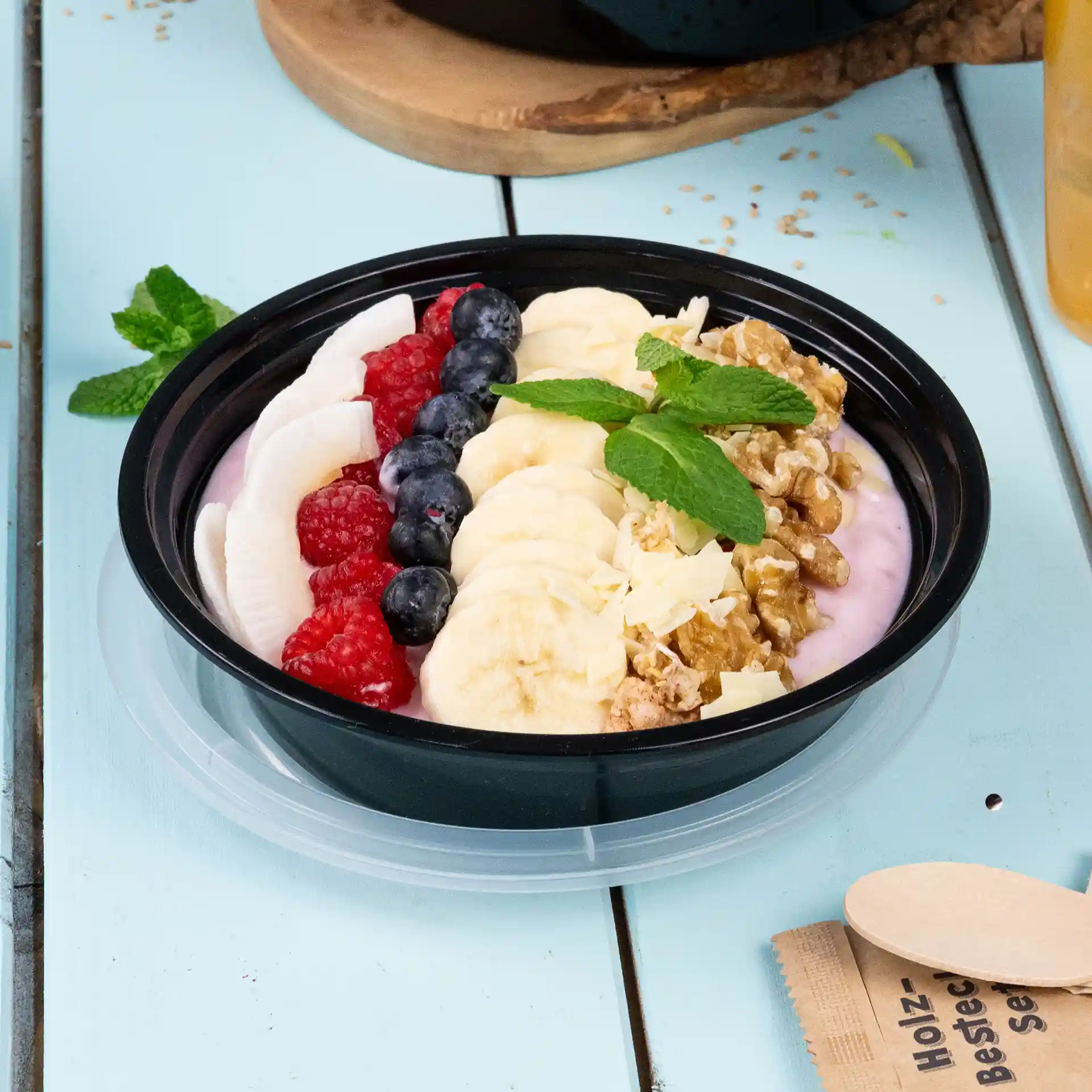 Assiette ronde noire avec du yaourt, des tranches de banane, des myrtilles, des framboises, du granola et des feuilles de menthe, sur une surface bleu clair.