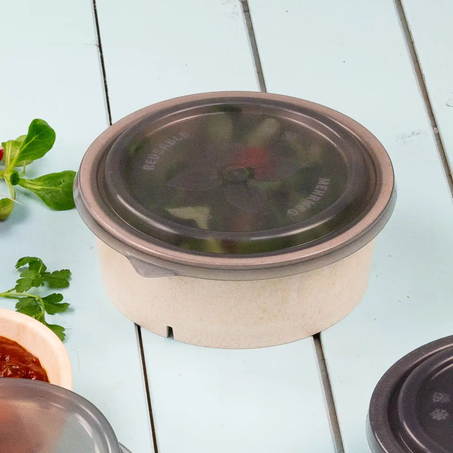 Bol rond avec un couvercle réutilisable gris, posé sur une surface bleu clair avec des herbes et de la sauce à proximité.