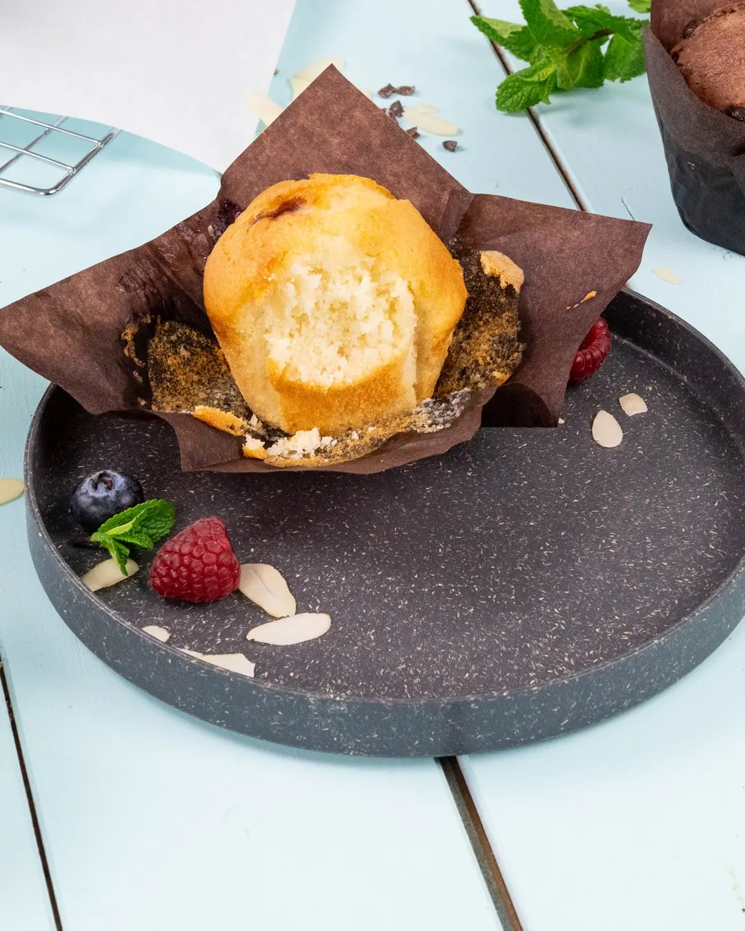 Muffin dans un emballage en papier brun sur une assiette sombre, entouré de framboises, myrtilles, feuilles de menthe et tranches d'amande.