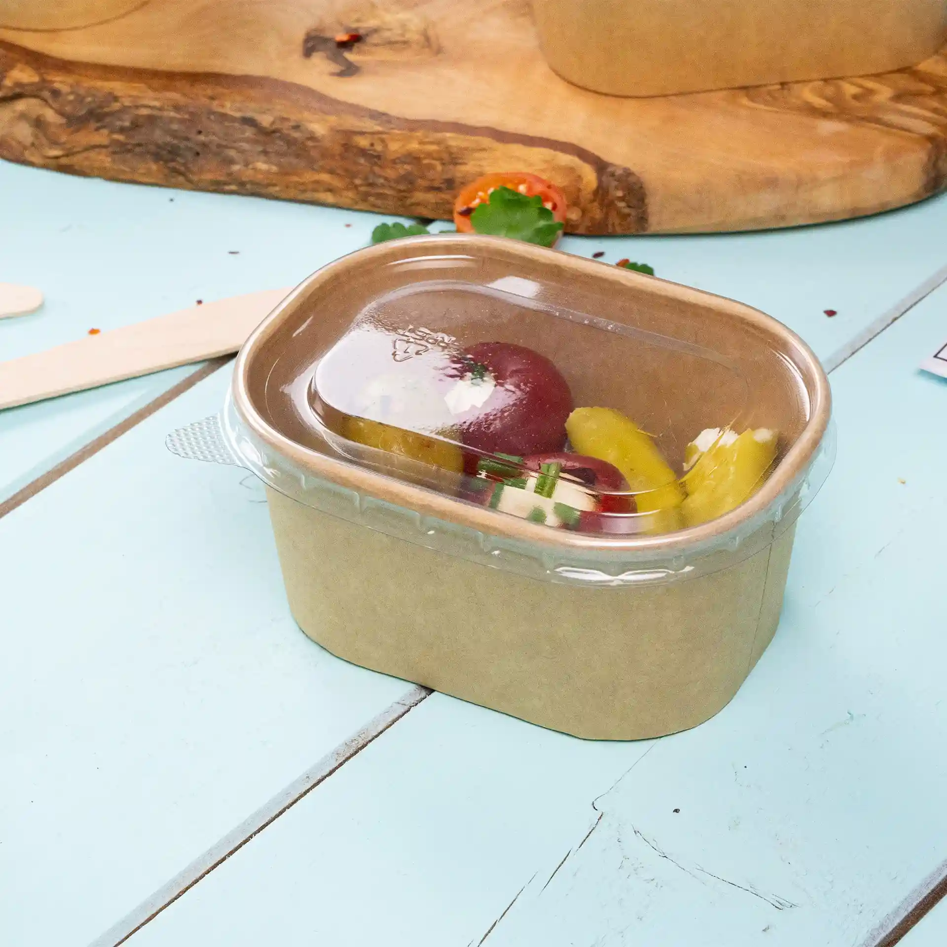 Conteneur alimentaire en kraft rectangulaire avec un couvercle transparent, contenant des légumes assortis, sur une surface bleu clair.