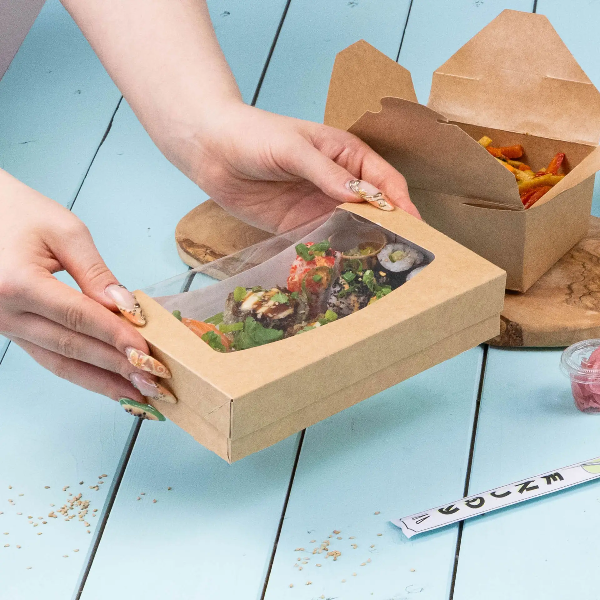 Une boîte en carton avec fenêtre contenant un sandwich au fromage frais, épinards et confiture de fruits, placée sur une surface teal.