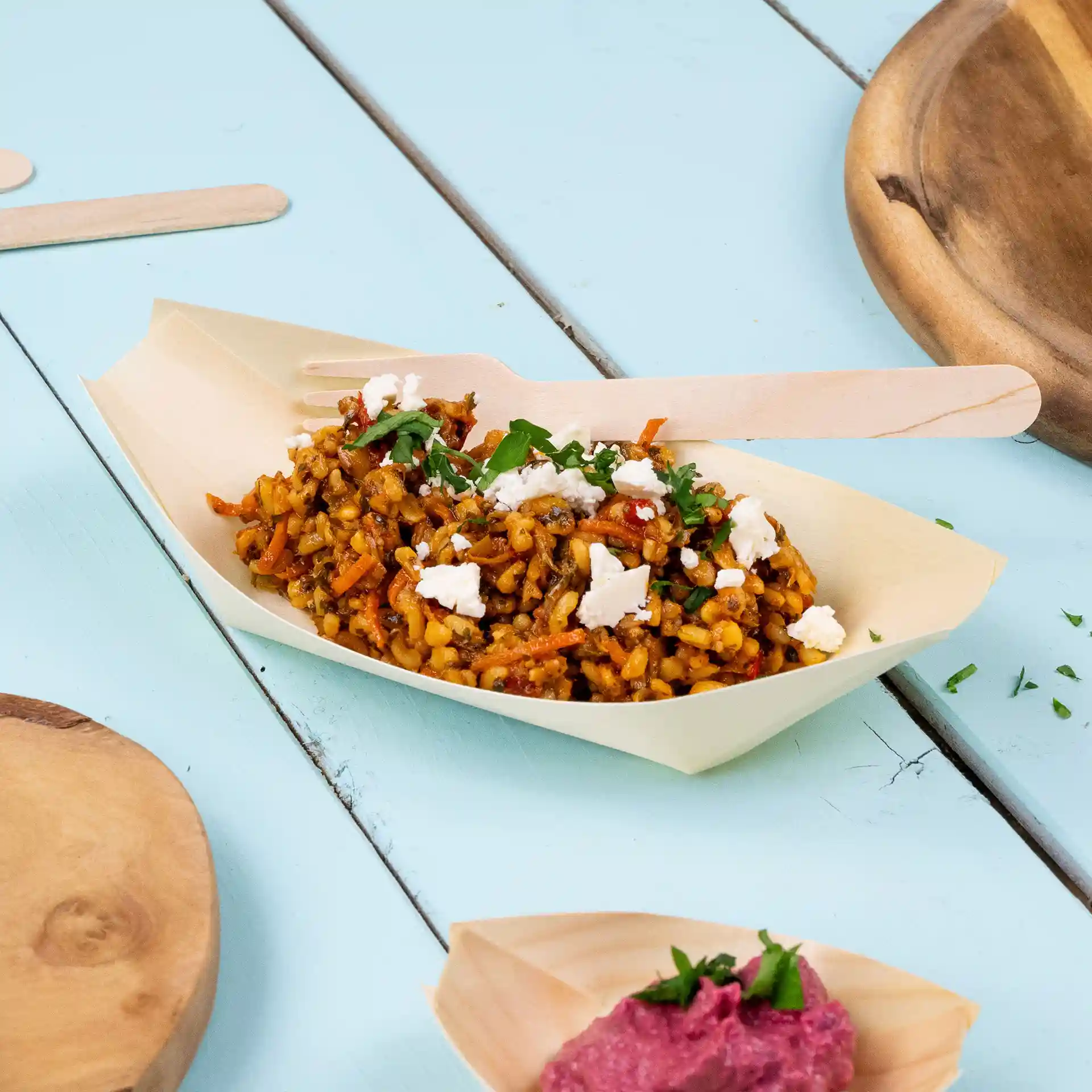 Plat en forme de bateau en bois avec du riz, des légumes et du fromage feta, accompagné d'une fourchette en bois sur une surface bleue.