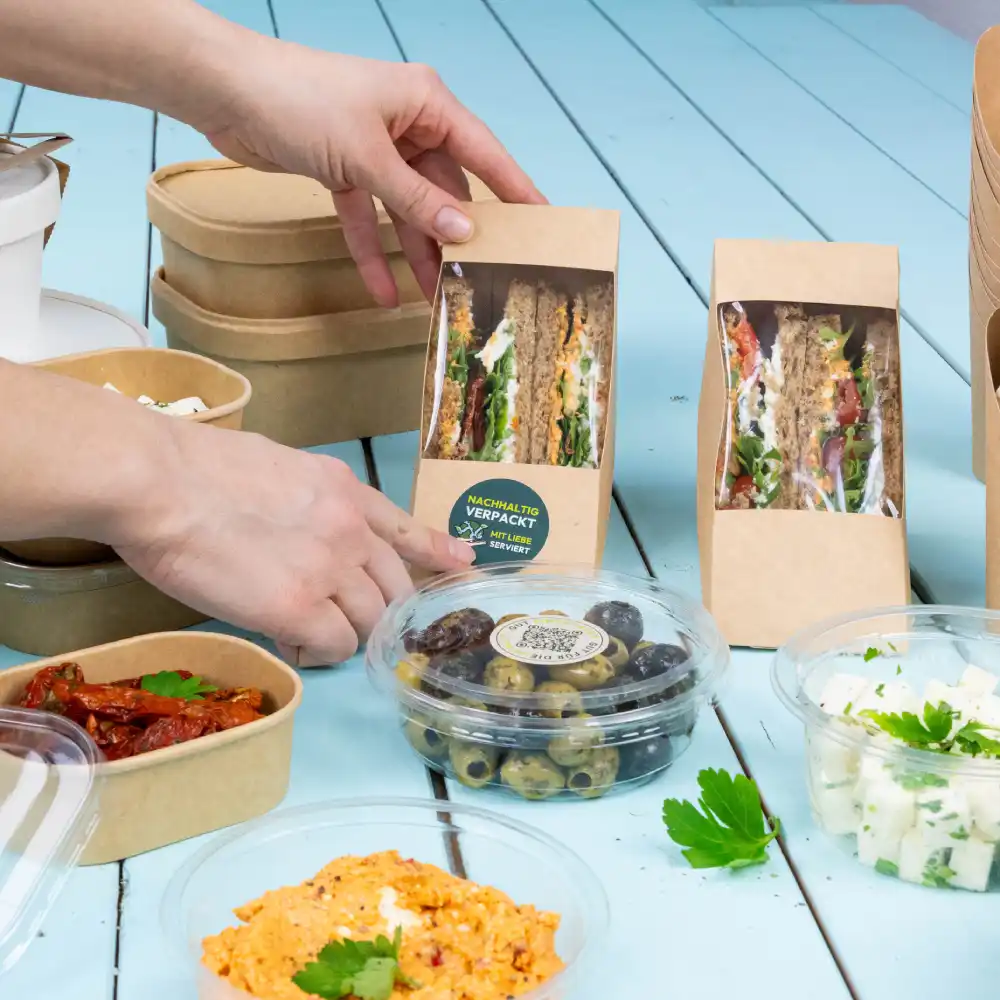 Une main tendue vers un paquet de sandwich sur une table ; divers contenants alimentaires, y compris des olives, un plat crémeux et des tomates séchées au soleil, sont visibles.