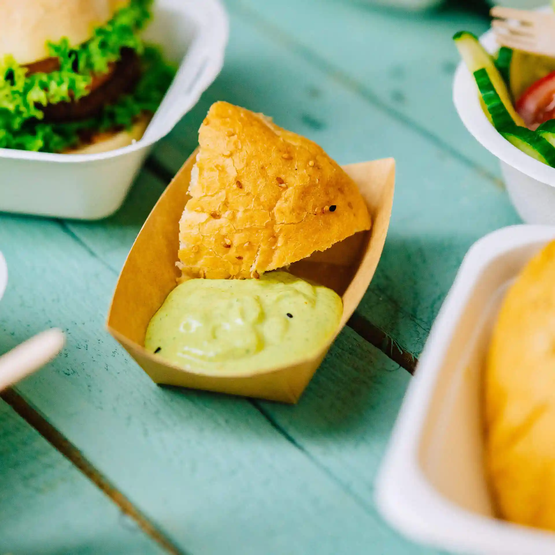 Plateau à snacks en carton brun contenant un morceau de pain frit et une sauce verte sur une surface en bois bleu.