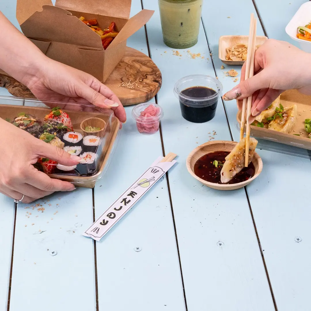Des mains tenant des sushis dans un récipient transparent, avec de la sauce, du gingembre mariné et des baguettes sur une table bleue.
