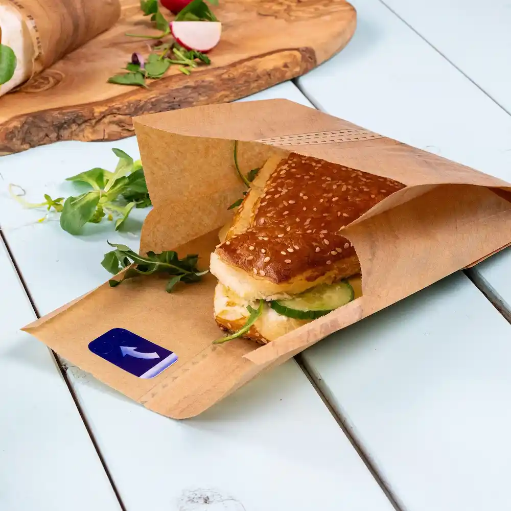 Sac en papier brun contenant un sandwich aux graines de sésame avec des concombres, entouré de légumes frais sur une surface bleue.
