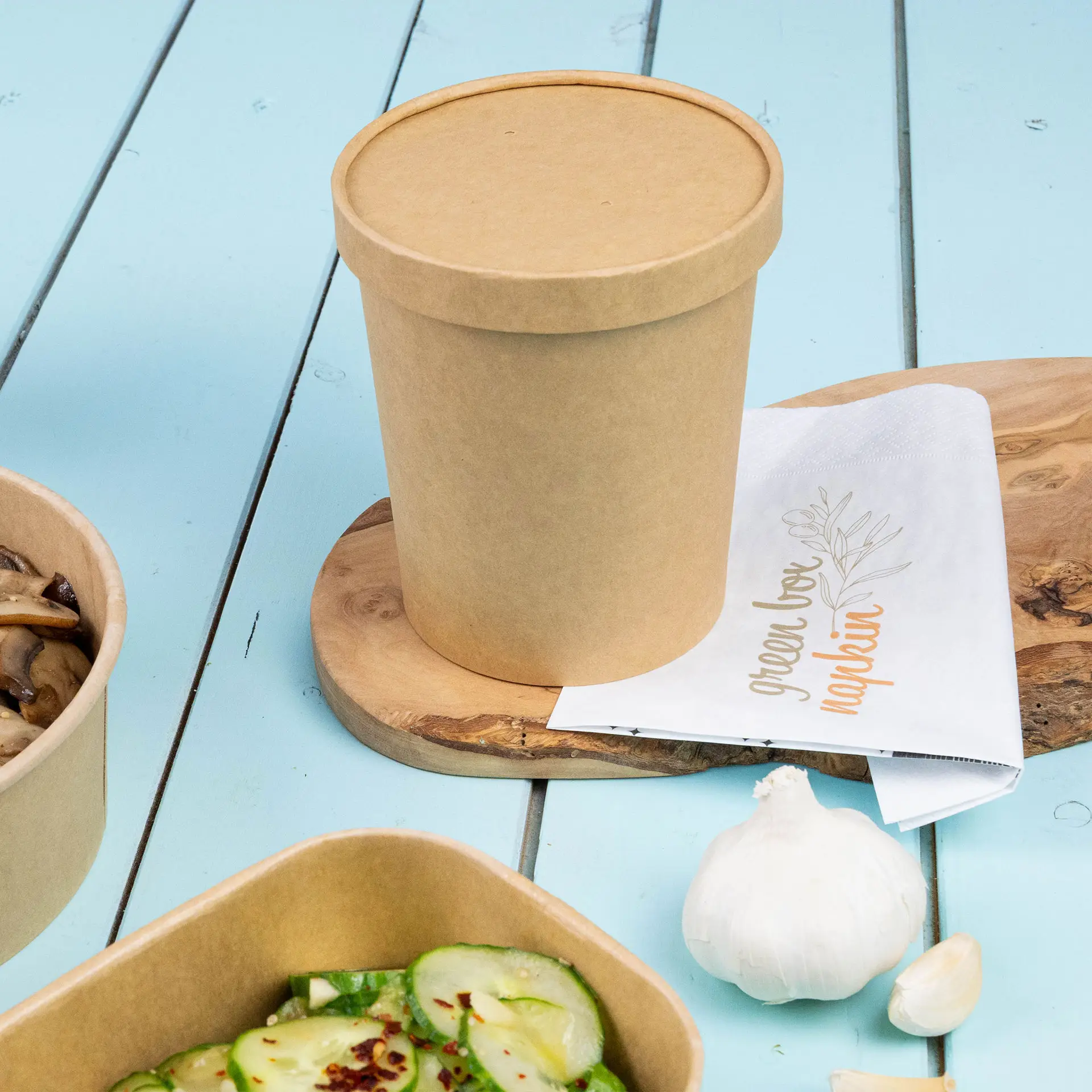 Gobelet en carton brun pour soupe avec couvercle, posé sur une planche en bois, à côté d'une serviette et de contenants alimentaires.