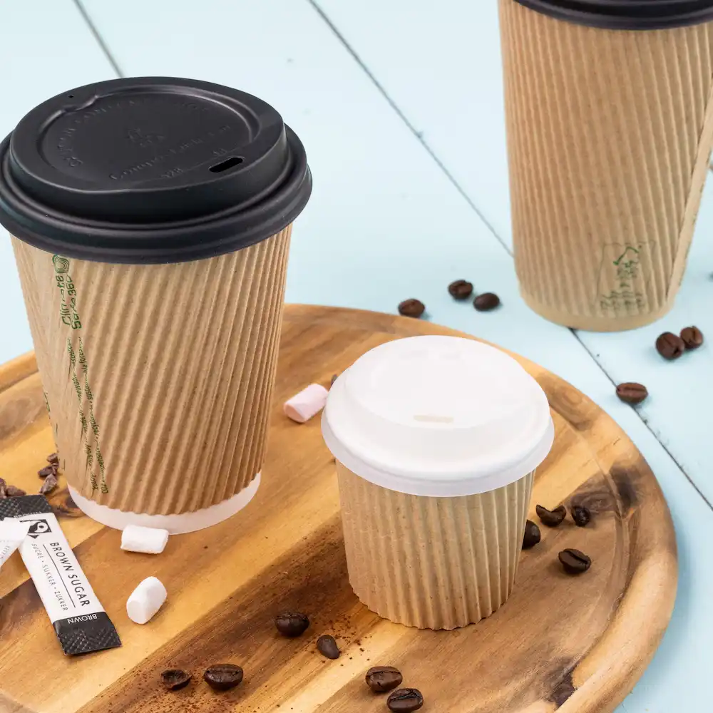 Trois gobelets en papier de tailles variées avec couvercles, placés sur un plateau en bois avec des grains de café et des sachets de sucre.