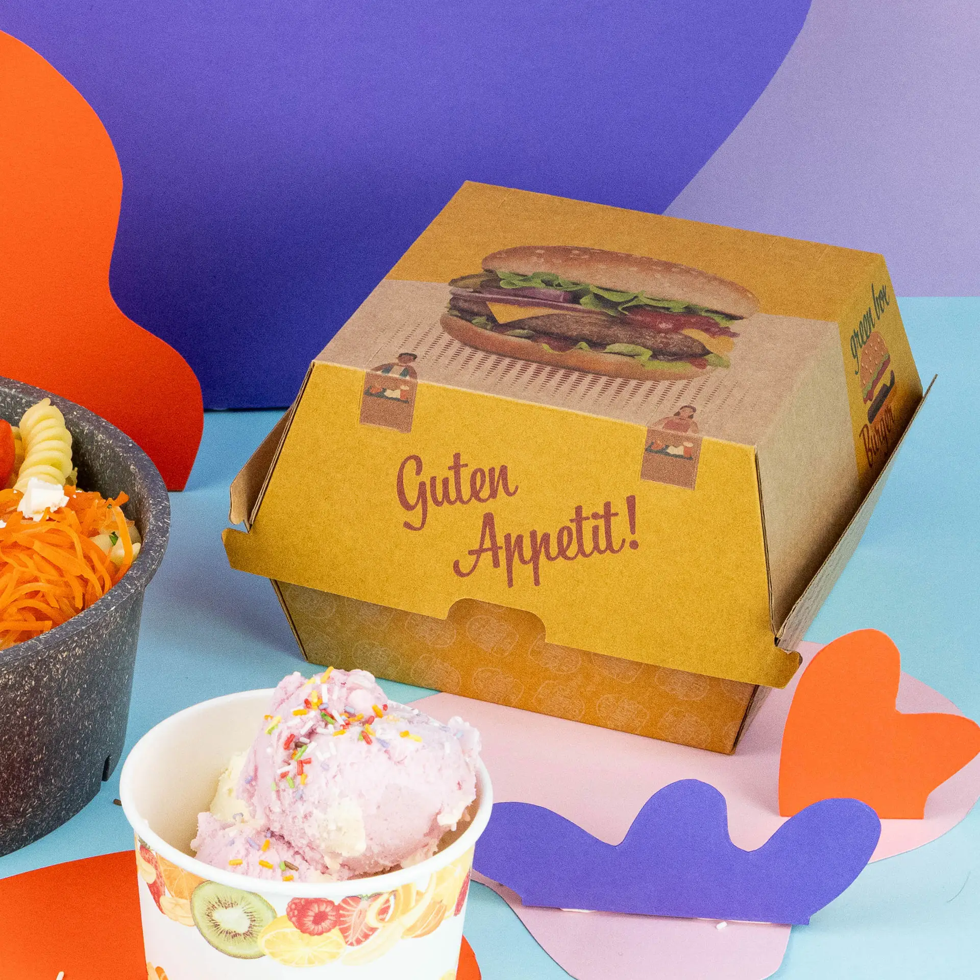 Un burger entier avec des graines de sésame dans une boîte en carton kraft, à côté d'un bol de légumes mélangés et d'un petit contenant de sauce.