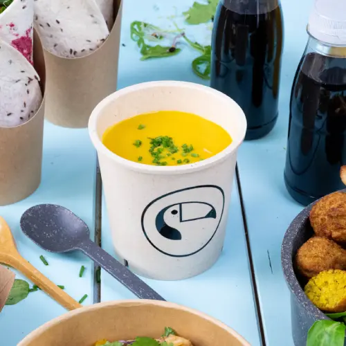 Bol de soupe avec de la soupe jaune et des garnitures vertes, entouré de contenants alimentaires et de boissons sur une surface bleue.