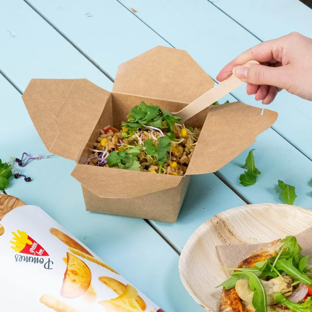 Boîte à emporter marron remplie de légumes mélangés et d'herbes, avec une main tenant une cuillère en bois, sur une table bleue.