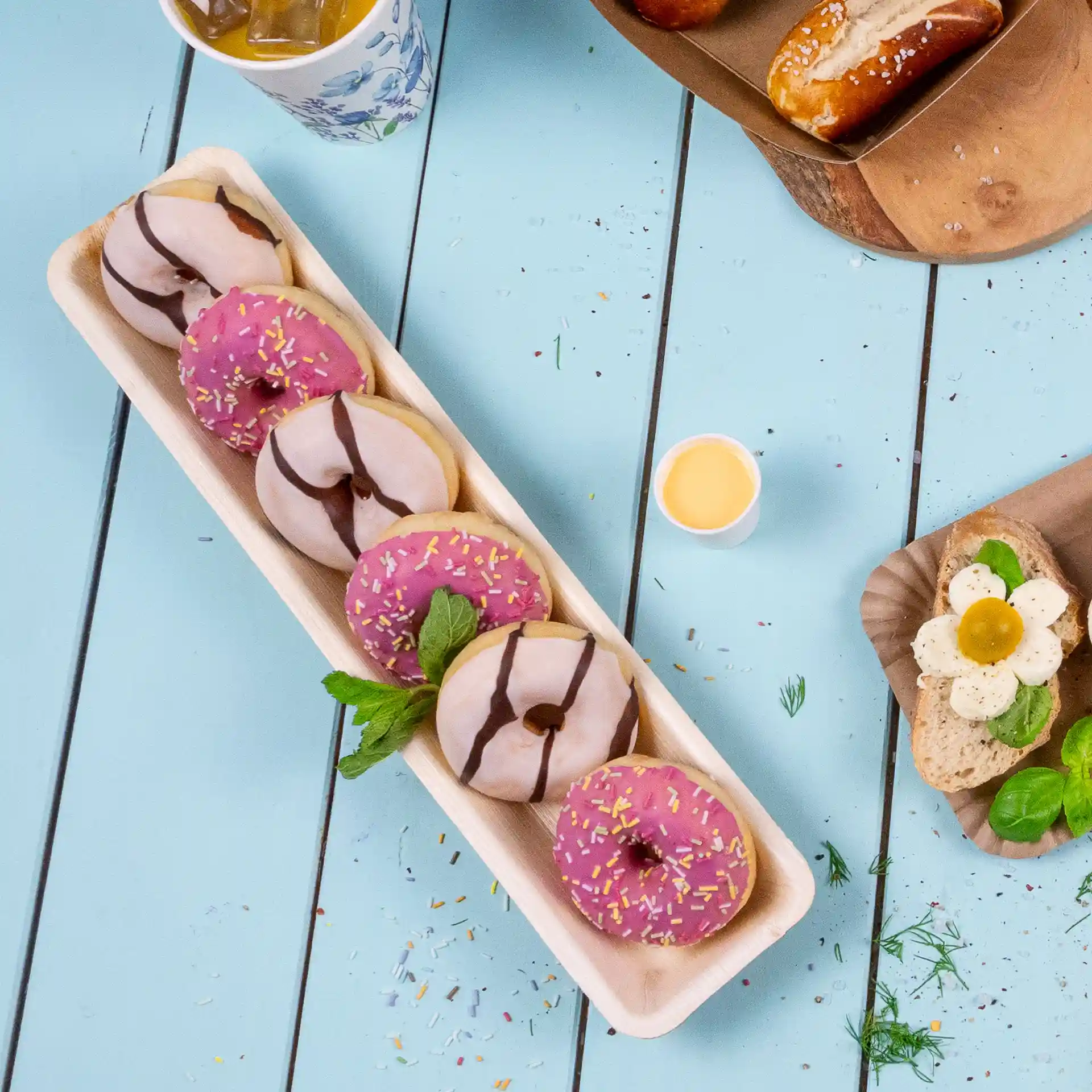 Plateau rectangulaire en feuille de palmier avec plusieurs beignets, y compris des variétés roses et chocolat, sur une surface en bois bleu.