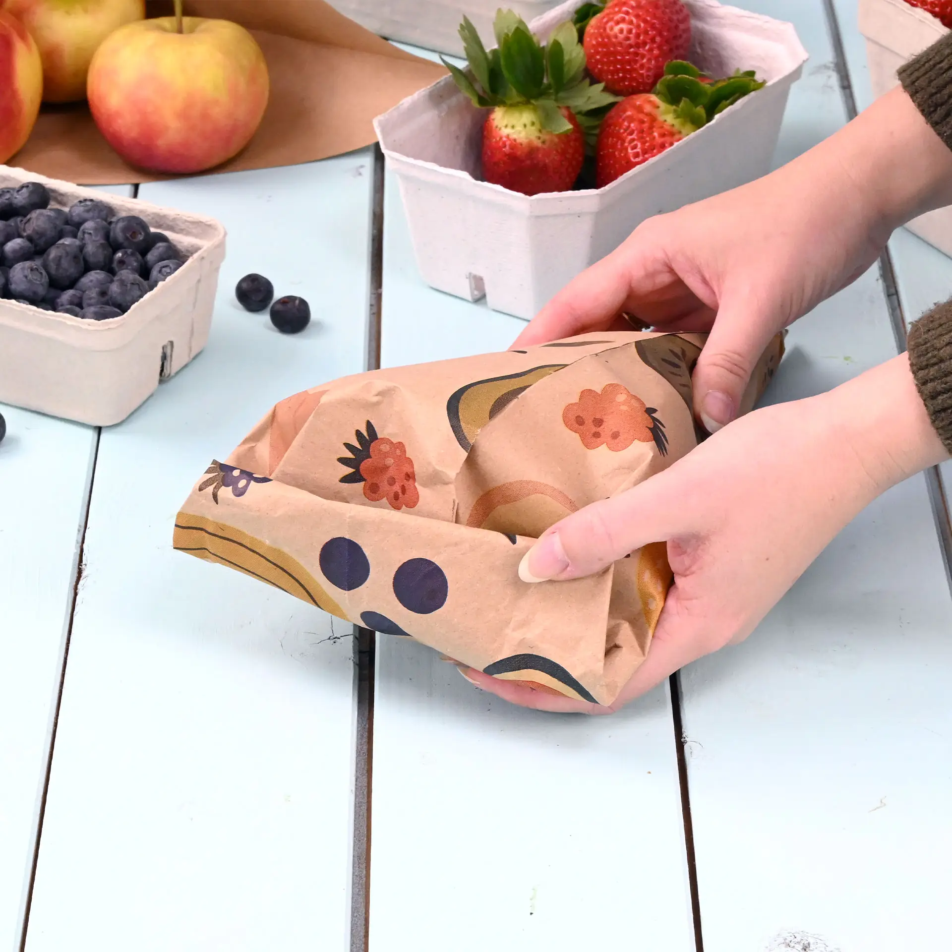 Des mains tenant un cône en papier imprimé personnalisé avec un design brun, entouré de corbeilles de fruits sur une table en bois.