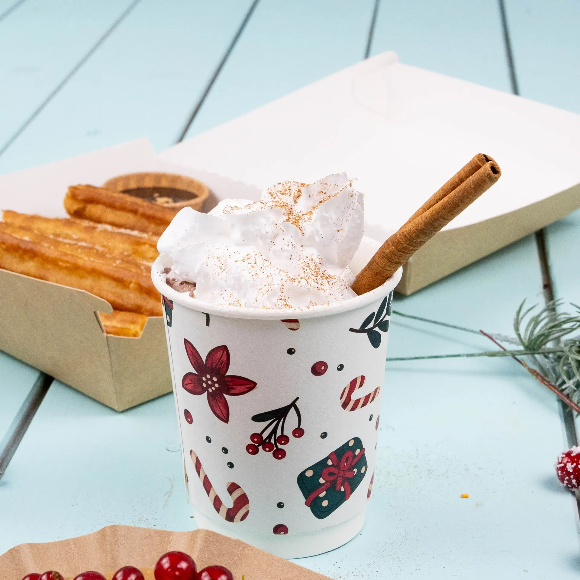 Tasse double paroi avec design d'hiver, remplie de crème fouettée et d'un bâton de cannelle, à côté de churros dans une boîte.