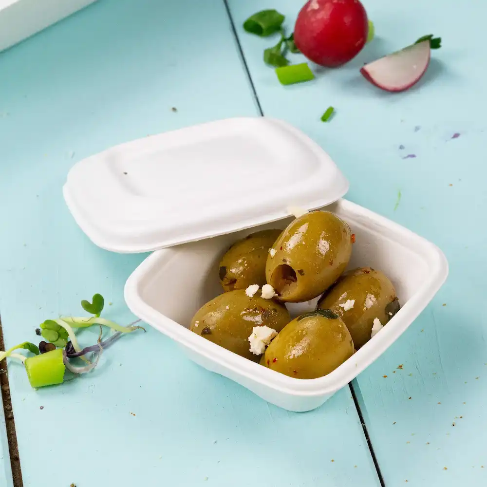 Contenant avec plusieurs olives vertes garnies, sur un fond bleu clair, avec un radis et des herbes à côté.