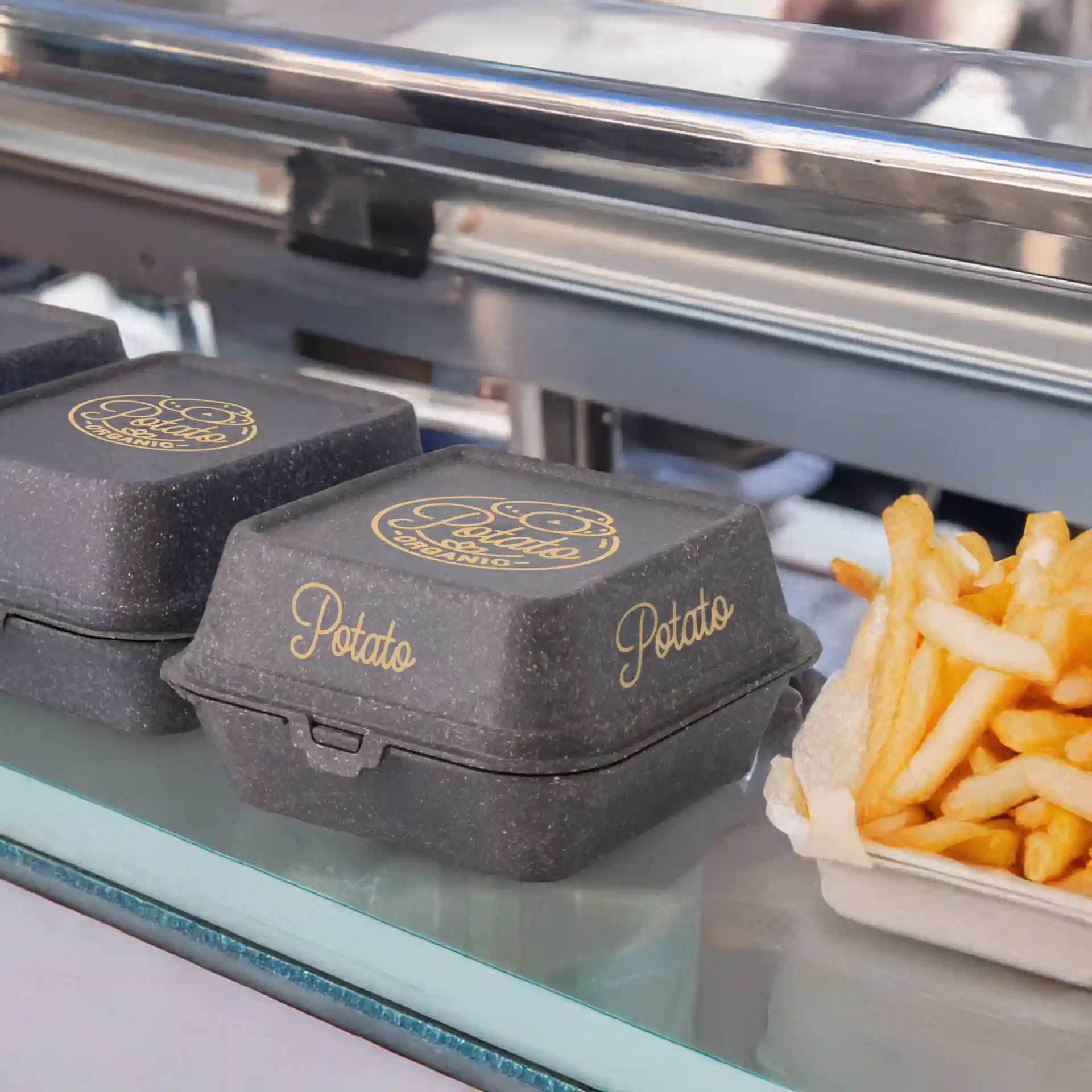 Boîtes à burgers réutilisables grises imprimées en forme rectangulaire, présentées avec des frites sur une surface en verre.