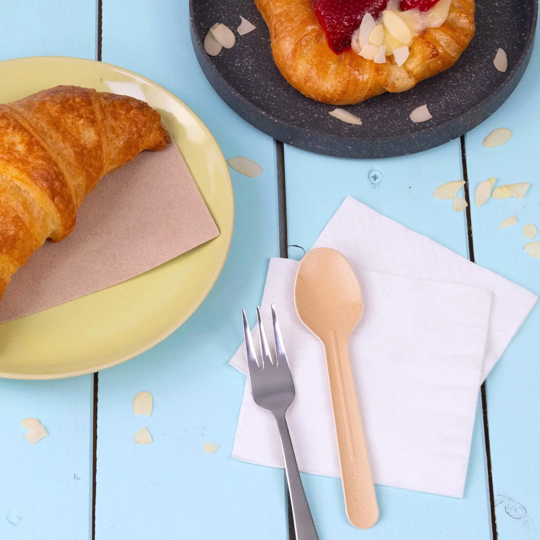 Image d'un croissant sur une assiette jaune, une pâtisserie sur une assiette sombre, une cuillère en papier, une fourchette et des serviettes sur une table en bois bleue.