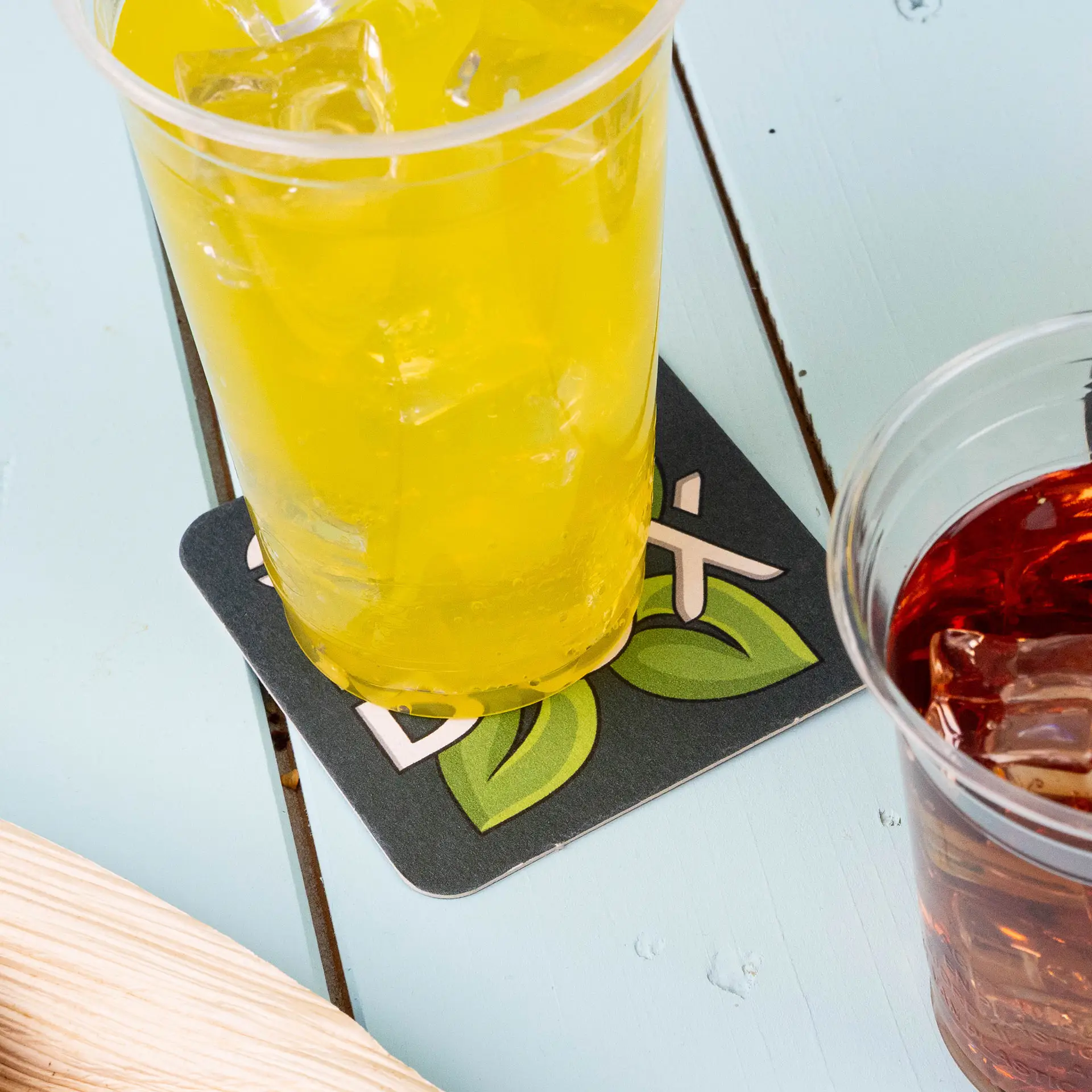 Une boisson jaune dans un gobelet en plastique repose sur un dessous de verre en papier carré imprimé avec des feuilles et le mot 'Box'.