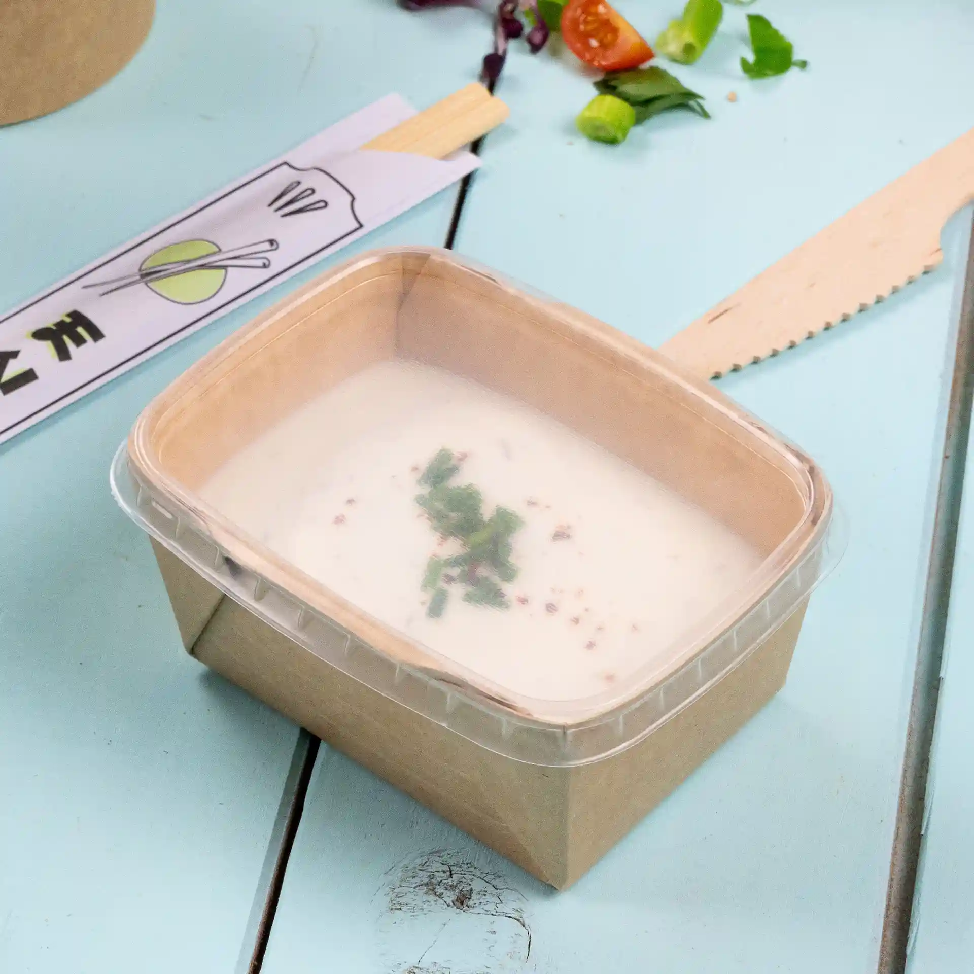 Gobelet rectangulaire transparent avec un couvercle clair, contenant une soupe crémeuse garnie d'herbes vertes.