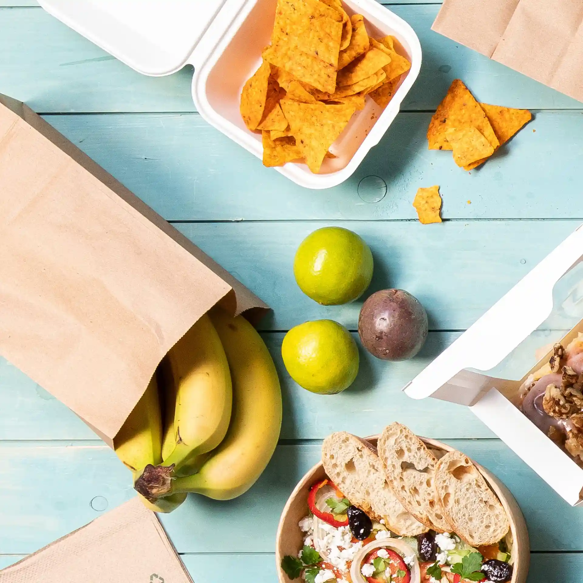 Image d'un sac en papier kraft marron avec des bananes, un bol de salade, des limes, un fruit de la passion et un contenant de chips sur une surface en bois bleu.