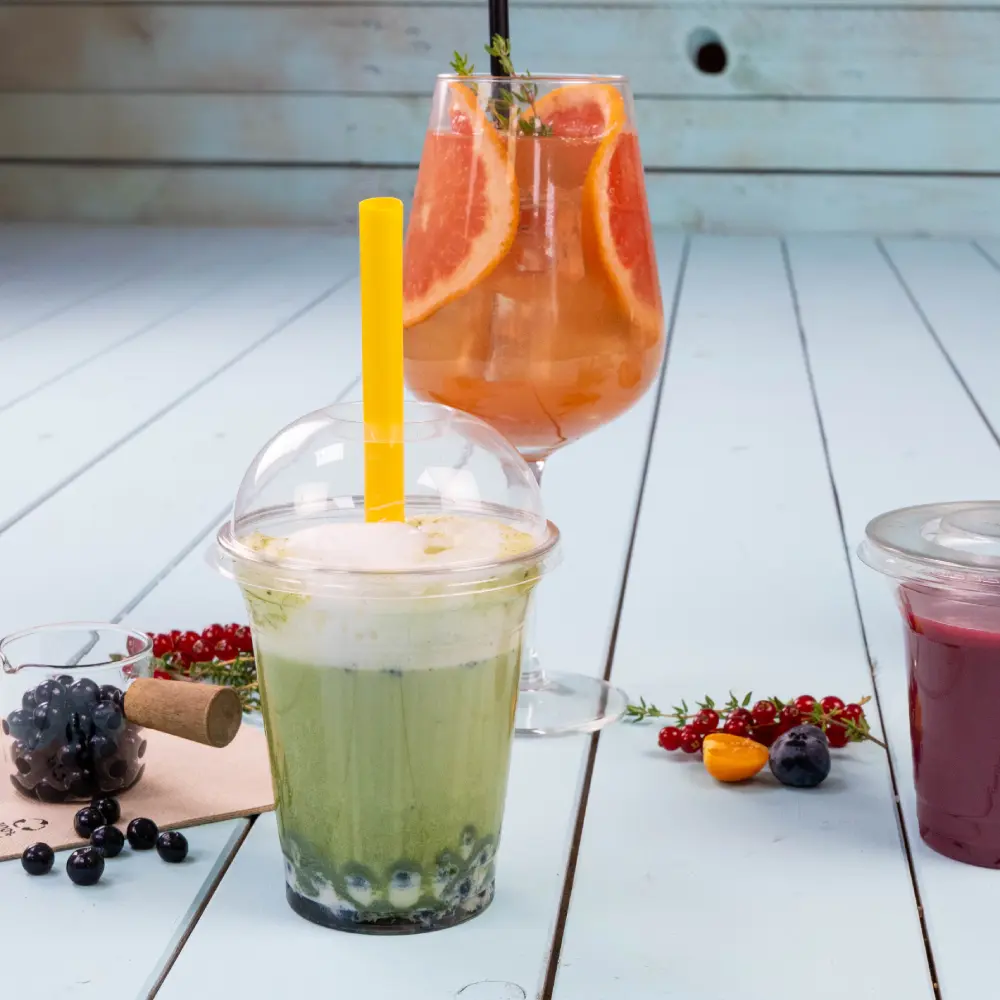 Une boisson verte avec une paille jaune dans un gobelet transparent, avec des perles de tapioca bleues au fond ; un cocktail coloré avec des tranches de pamplemousse dans un verre derrière.