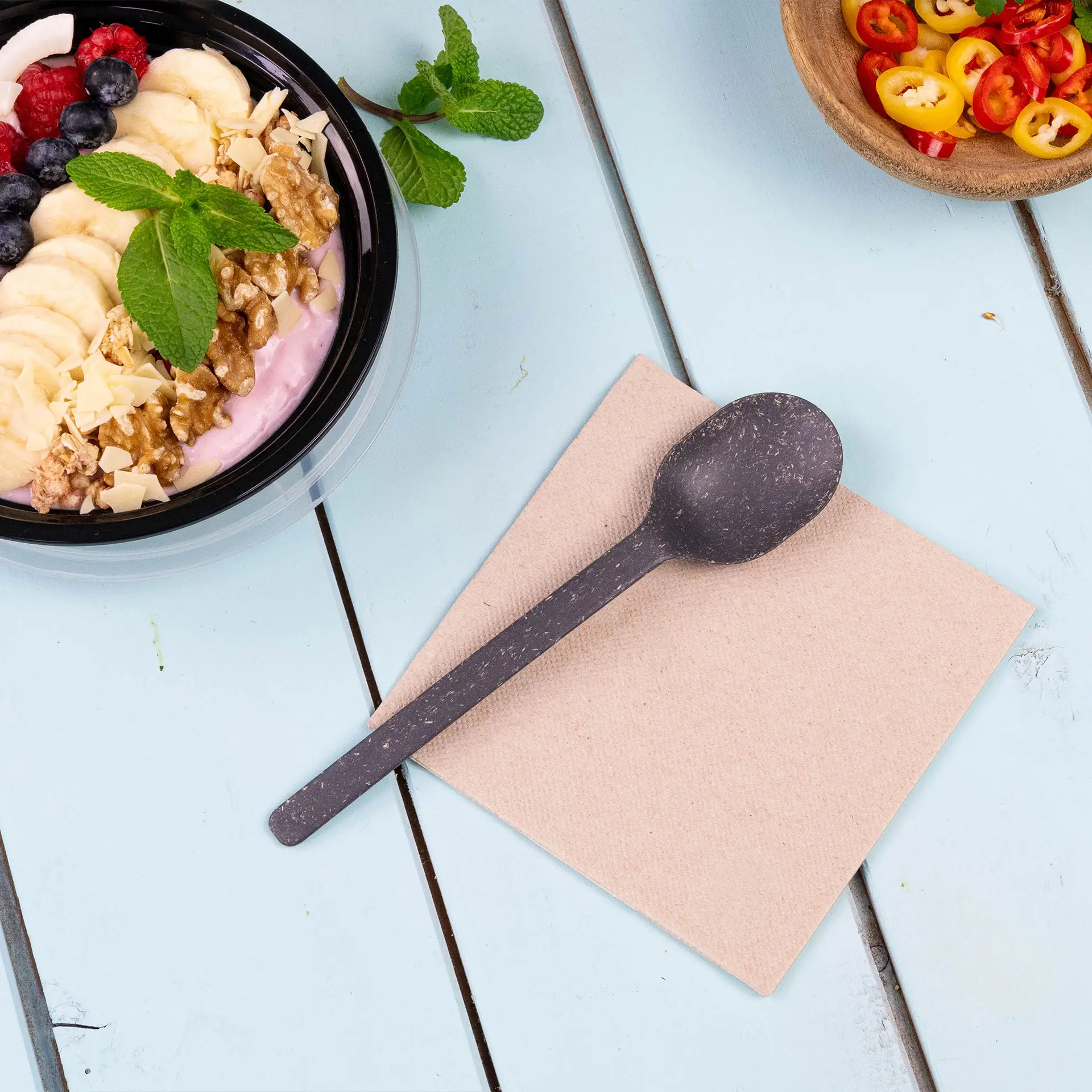 Image d'une cuillère noire sur une serviette beige, avec un bol de yaourt garni de fruits et de noix en arrière-plan.