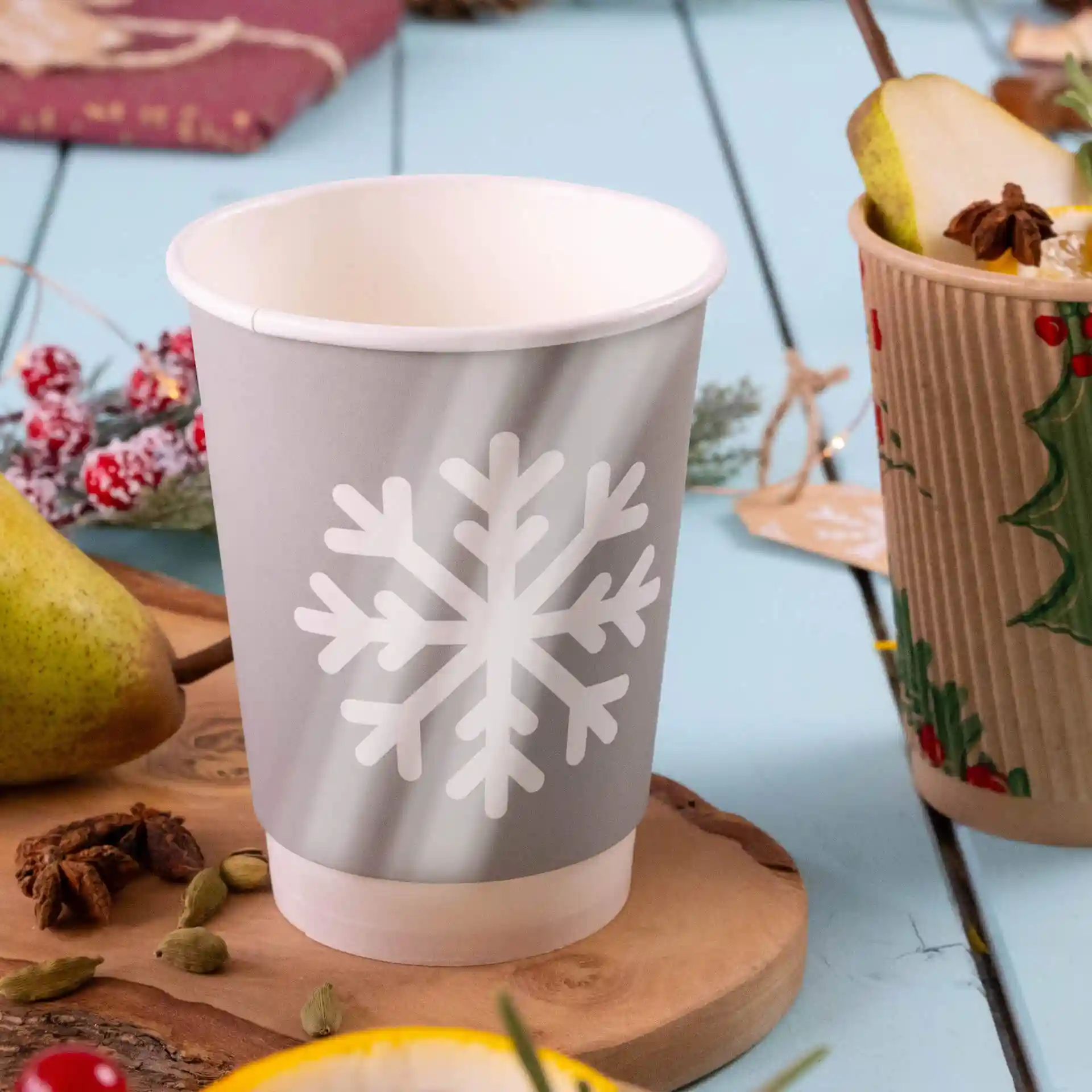 Gobelet en papier gris avec un design de flocon de neige blanc, double paroi, posé sur une surface en bois avec une poire et des épices à proximité.